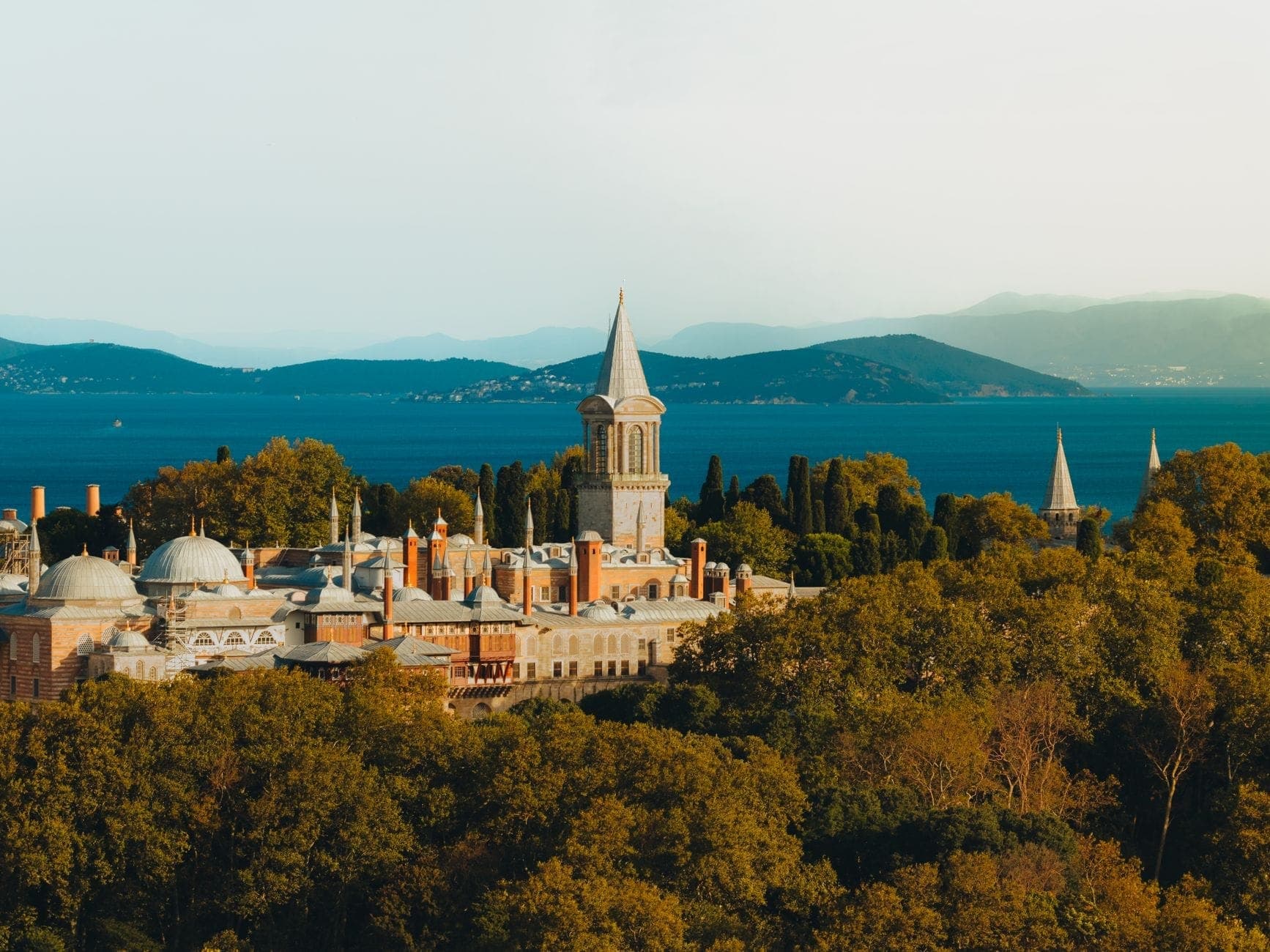 Ein Panoramablick auf den Topkapı-Palast mit Kuppeln und Turm, die über üppige Bäume ragen, vor dem glitzernden blauen Wasser und den fernen Hügeln Istanbuls.