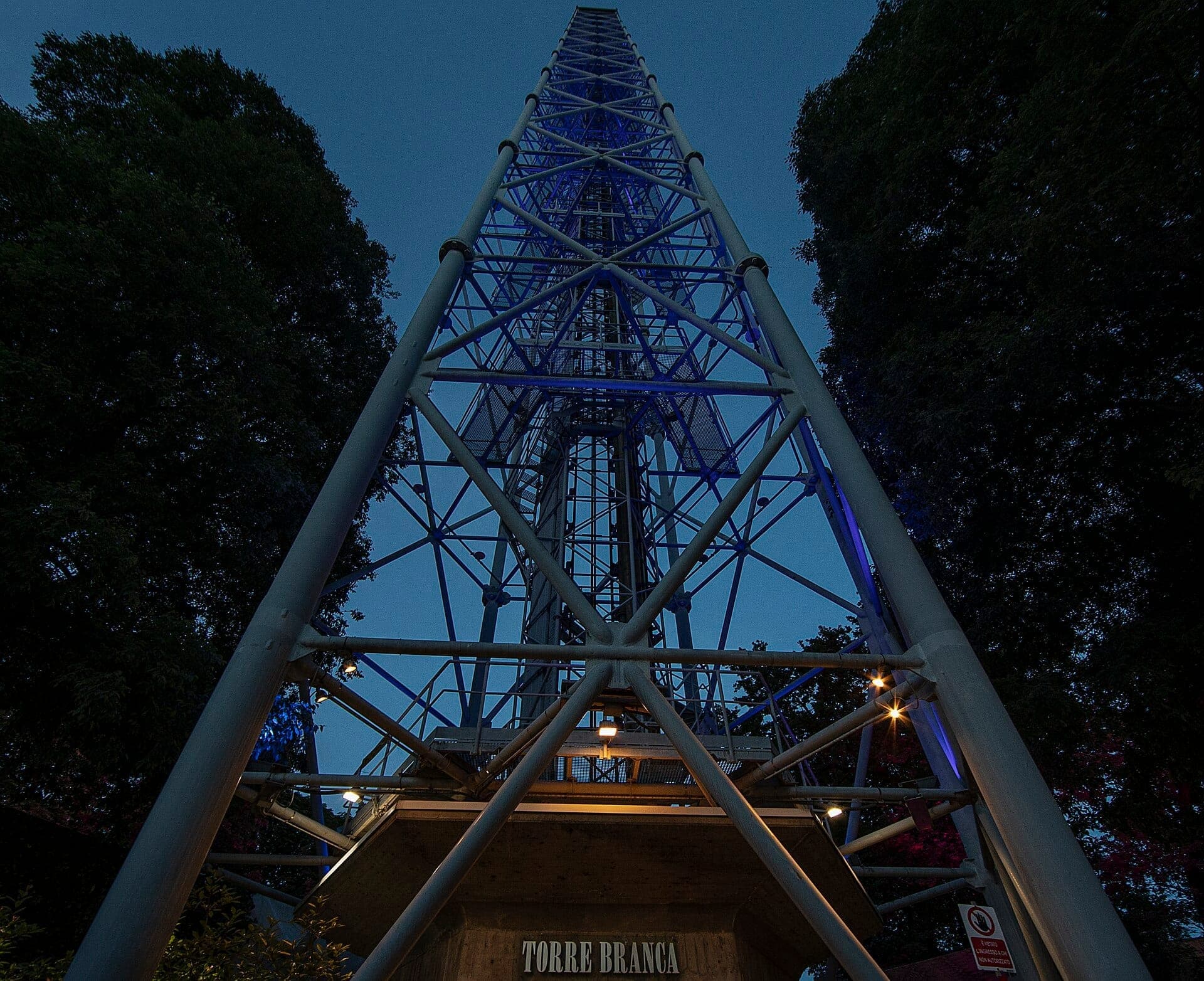 Base view of Torre Branca in Milan at dusk, with steel lattice structure lit by blue lights and framed by tall trees.