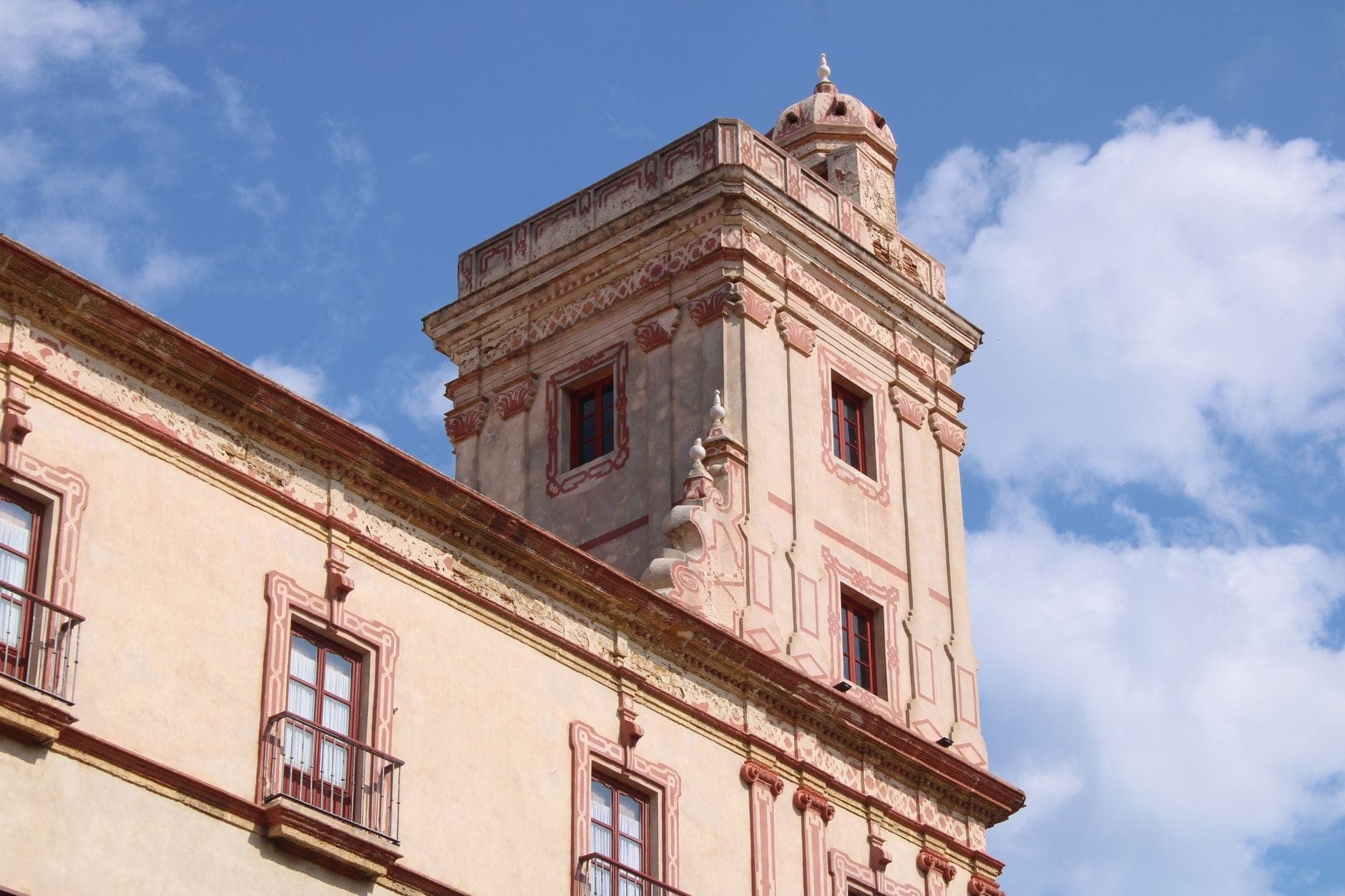 Tháp canh Torre Tavira vươn cao trên Cádiz, mặt tiền được trang trí tinh xảo và tháp hình chữ nhật tương phản với bầu trời xanh rực rỡ và những đám mây rải rác.