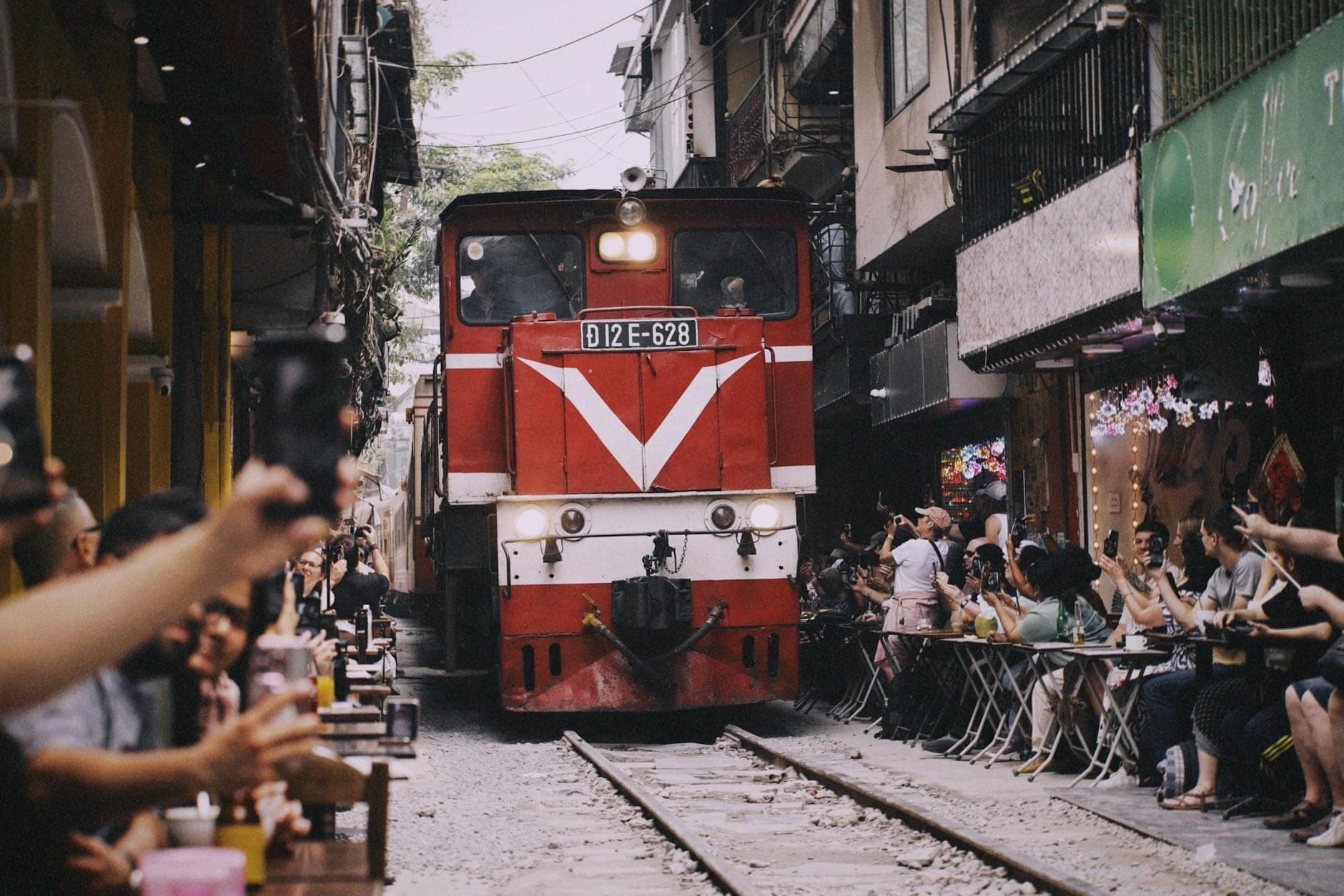 Một đoàn tàu đỏ chạy qua con ngõ dân sinh hẹp ở Hà Nội, hai bên là nhà cửa và quán cà phê ngoài trời với nhiều người đứng xem và chụp ảnh.