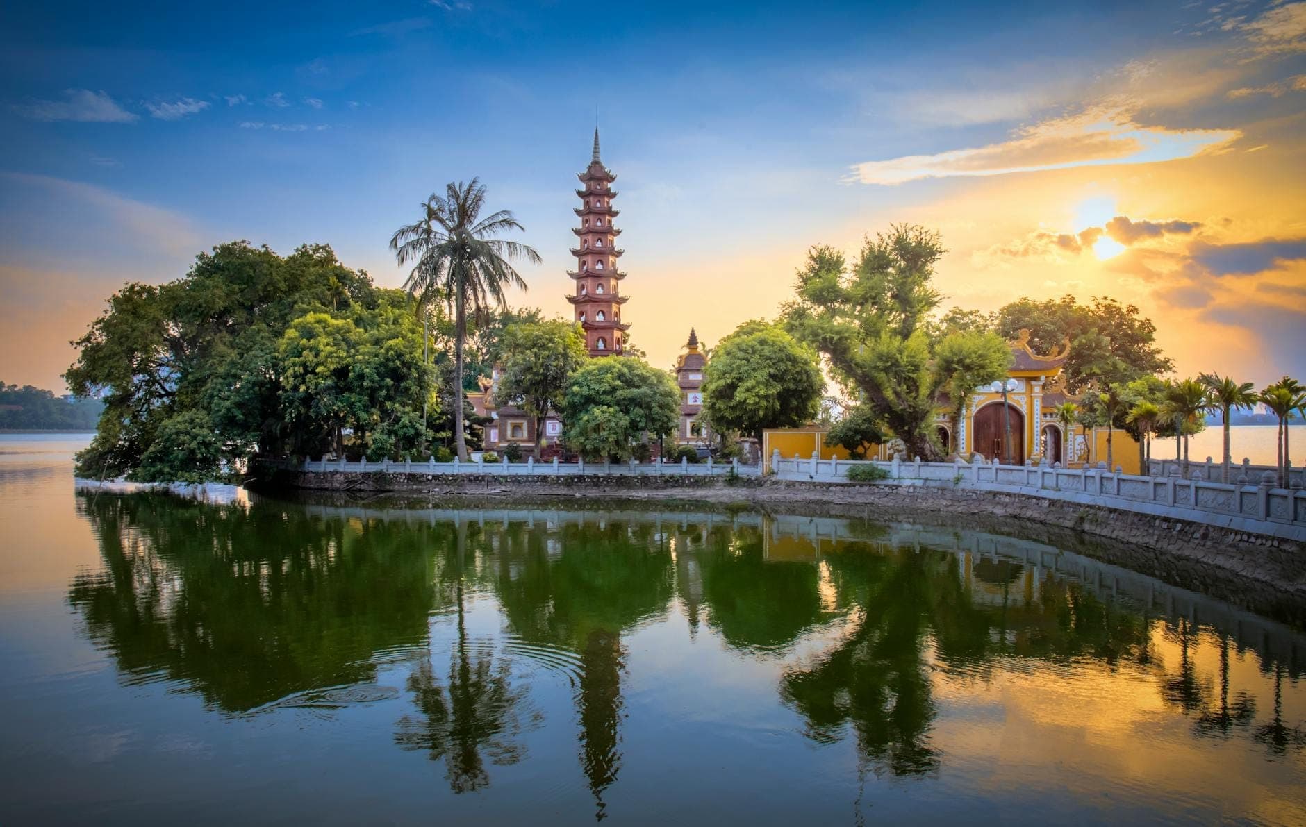 Chùa Trấn Quốc trên đảo nhỏ giữa Hồ Tây lúc hoàng hôn, cây xanh um tùm và mặt nước phẳng lặng phản chiếu hình ảnh ngôi chùa cổ kính, bầu trời rực rỡ và cổng tam quan.