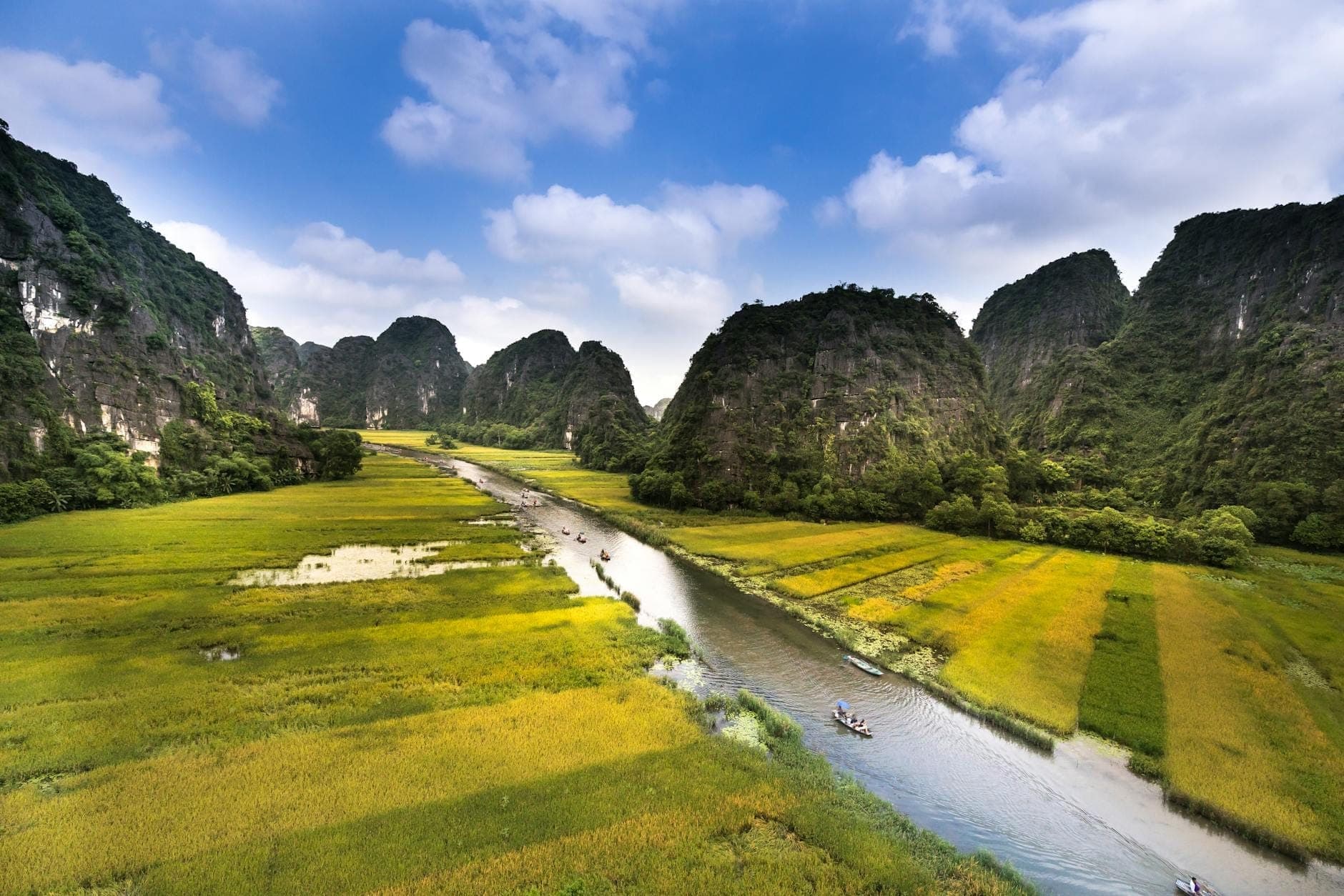 Toàn cảnh nhìn từ trên cao dòng sông Tràng An uốn lượn qua những cánh đồng xanh mướt và núi đá vôi hùng vĩ dưới bầu trời xanh điểm xuyết vài đám mây trắng.