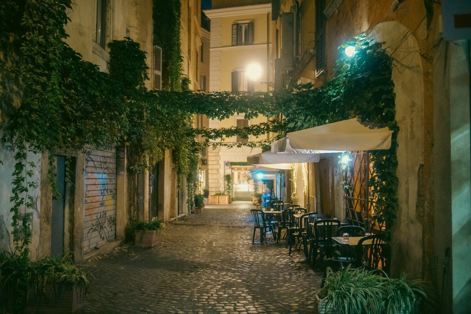 Tanawin sa gabi ng isang maganda at makitid na batong daanan sa Trastevere, na may mga gusaling may ibing at oker na kulay, mga mesa sa labas, at malambot na ilaw sa kalye.