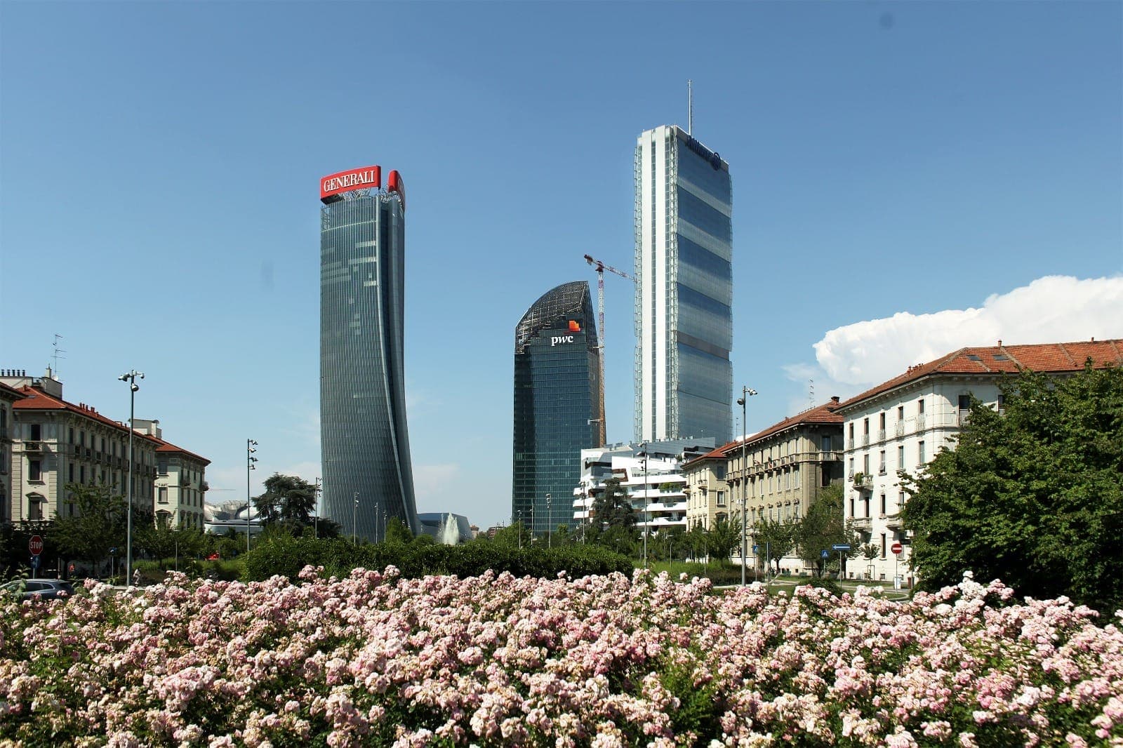 The Tre Torri skyscrapers in Milan’s CityLife district rise behind a blooming field of pink flowers, framed by traditional city buildings under a clear sky.