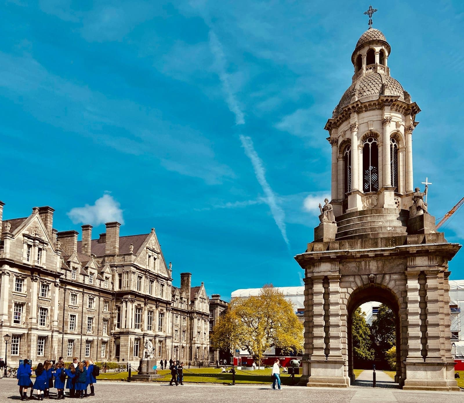 Innenhof des Trinity College Dublin mit dem ikonischen Campanile, historischen Steingebäuden, blauem Himmel und Besuchergruppen auf dem Gelände.