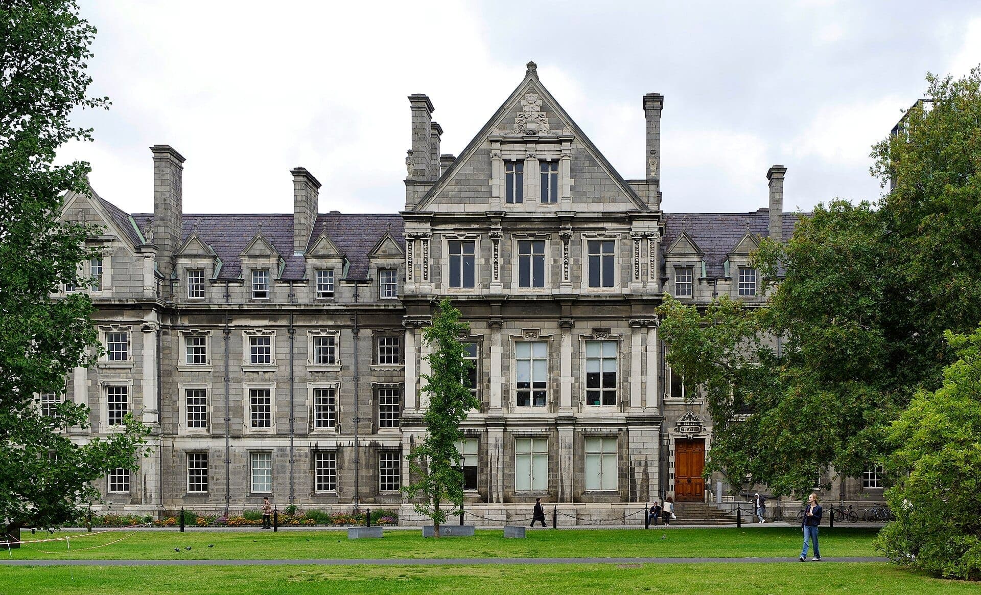 Fachada histórica de piedra del Trinity College Dublin con arquitectura georgiana, césped verde exuberante y visitantes paseando en un día nublado.