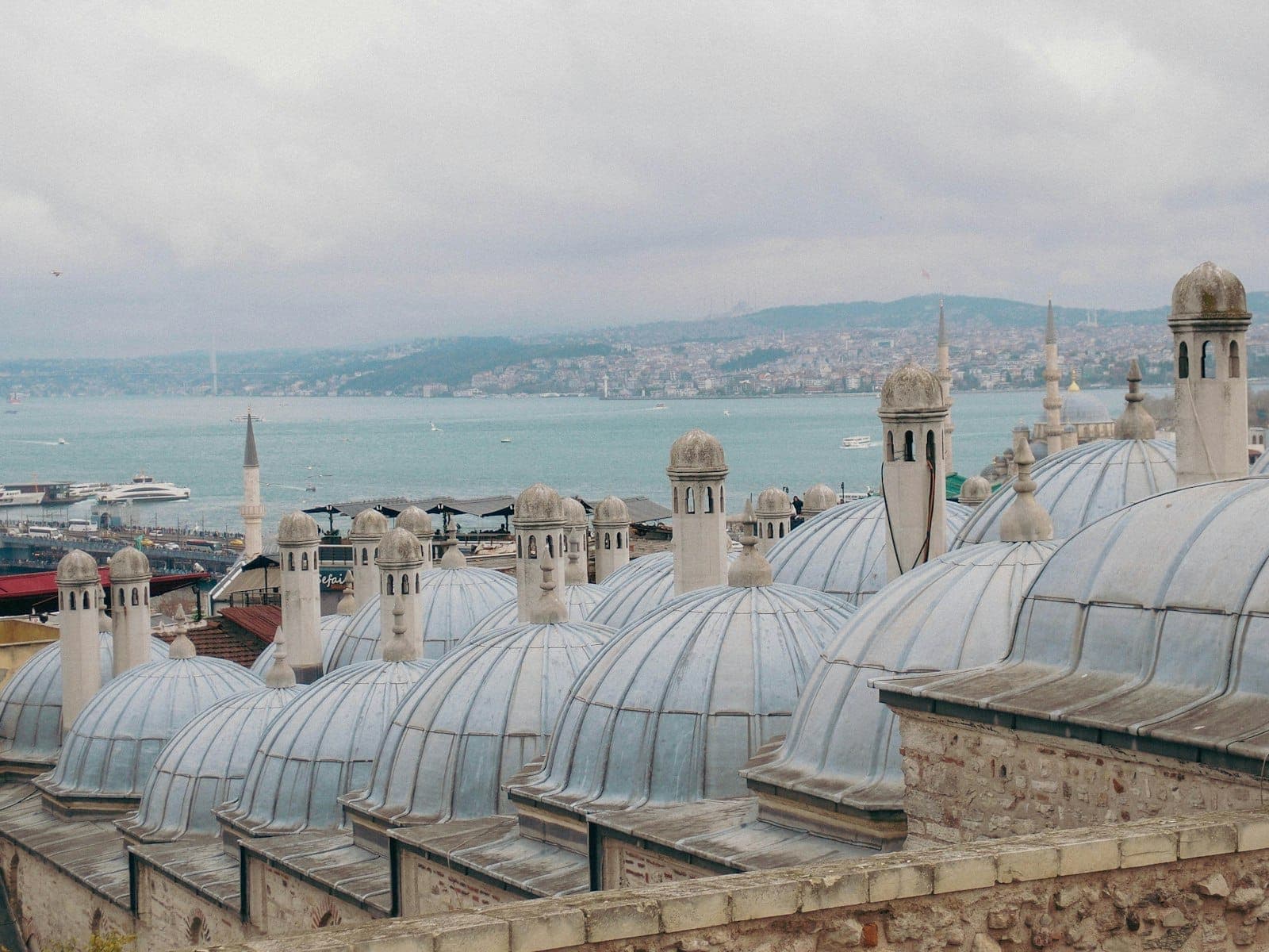 Kuppeln eines historischen Istanbuler Hamams auf dem Dach, im Hintergrund der Bosporus unter bewölktem Himmel.
