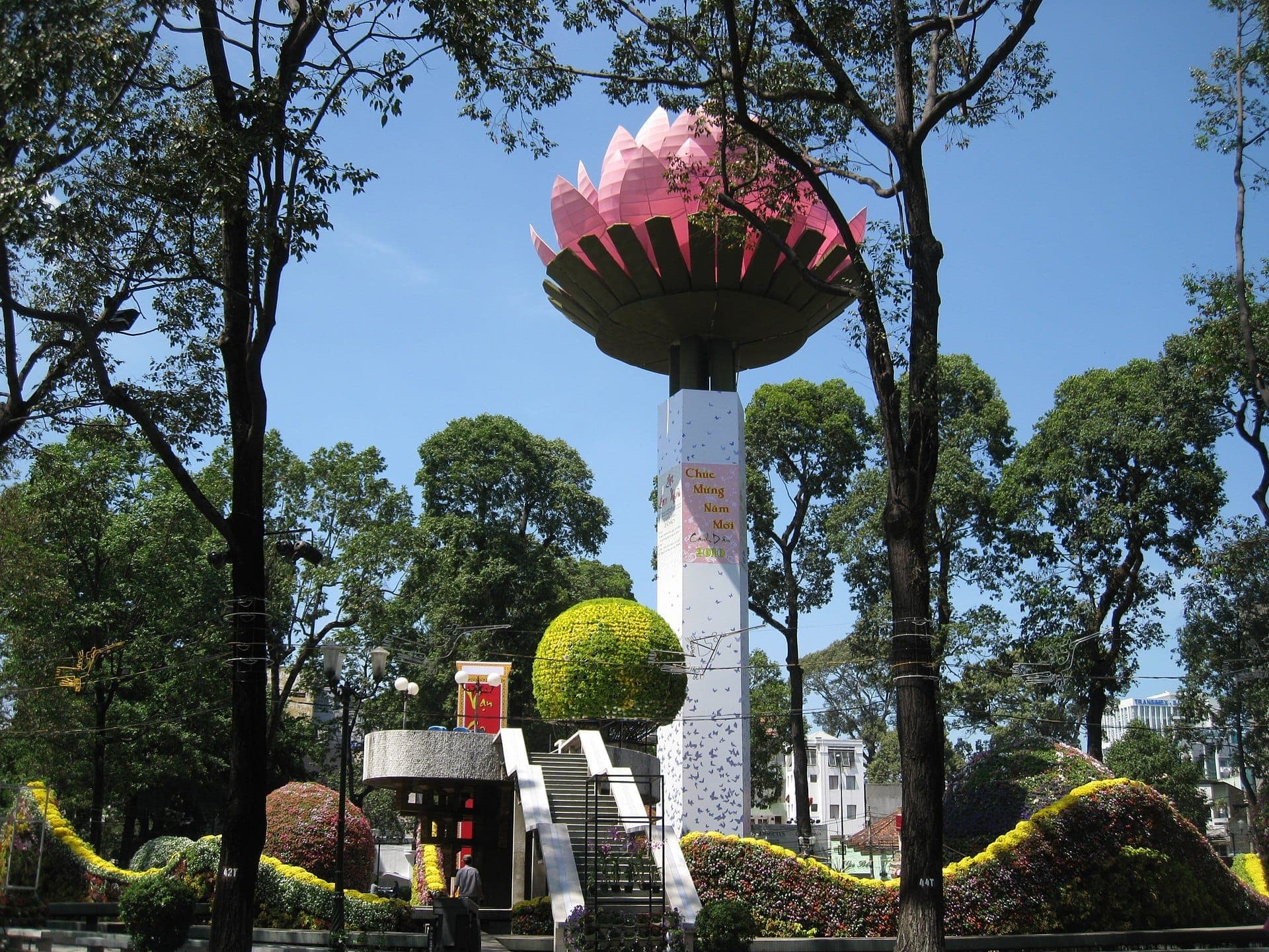 Vòng xuyến Turtle Lake nổi bật với cột bê tông cao gắn tượng hoa sen màu hồng, được bao quanh bởi cây cối xanh tươi và các chậu hoa rực rỡ dưới bầu trời xanh.