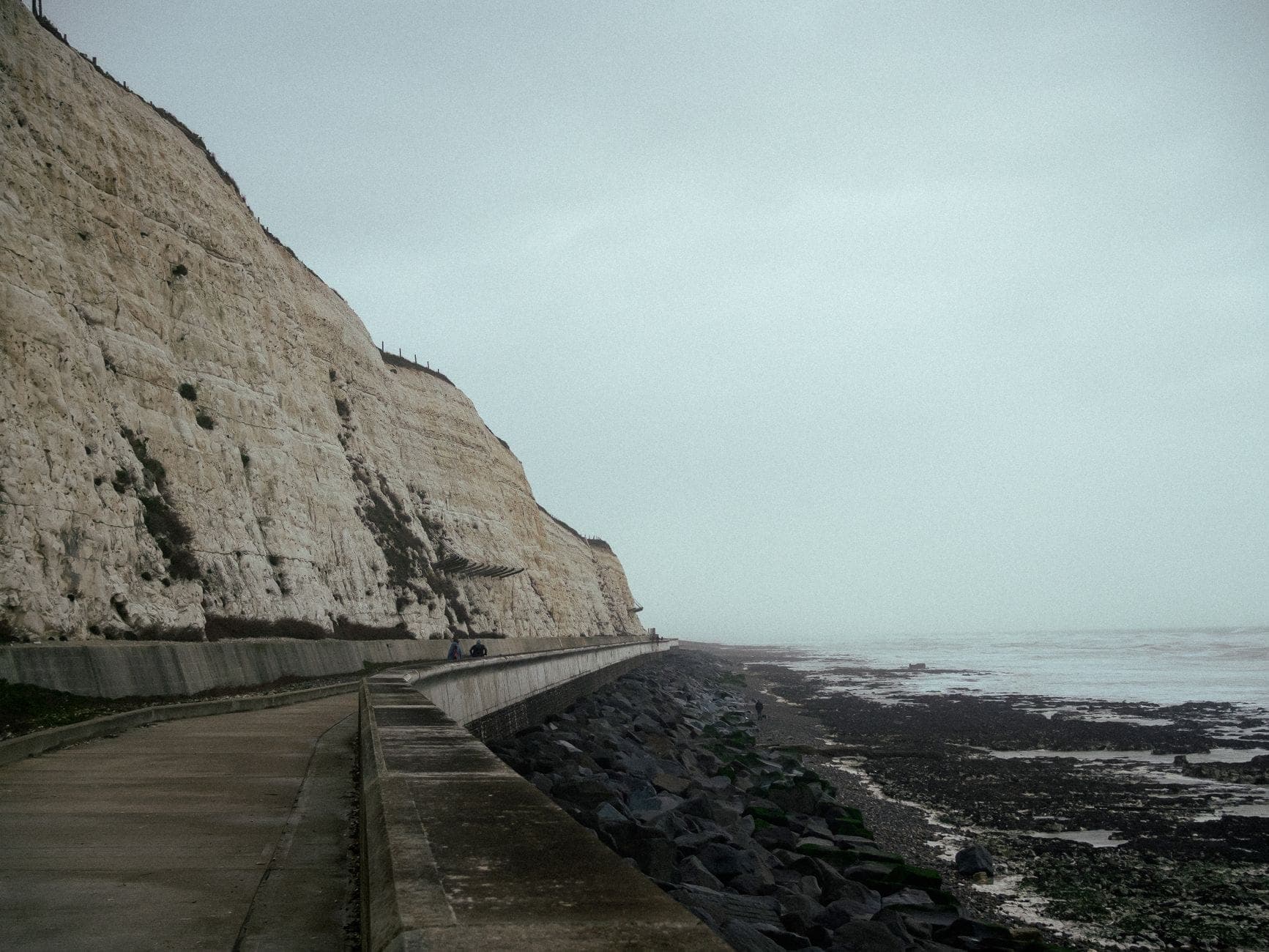 Lối đi bê tông Undercliff Walk Brighton uốn lượn dưới những vách đá phấn trắng cao vút dọc biển East Sussex, phía bên phải là biển dưới bầu trời xám.
