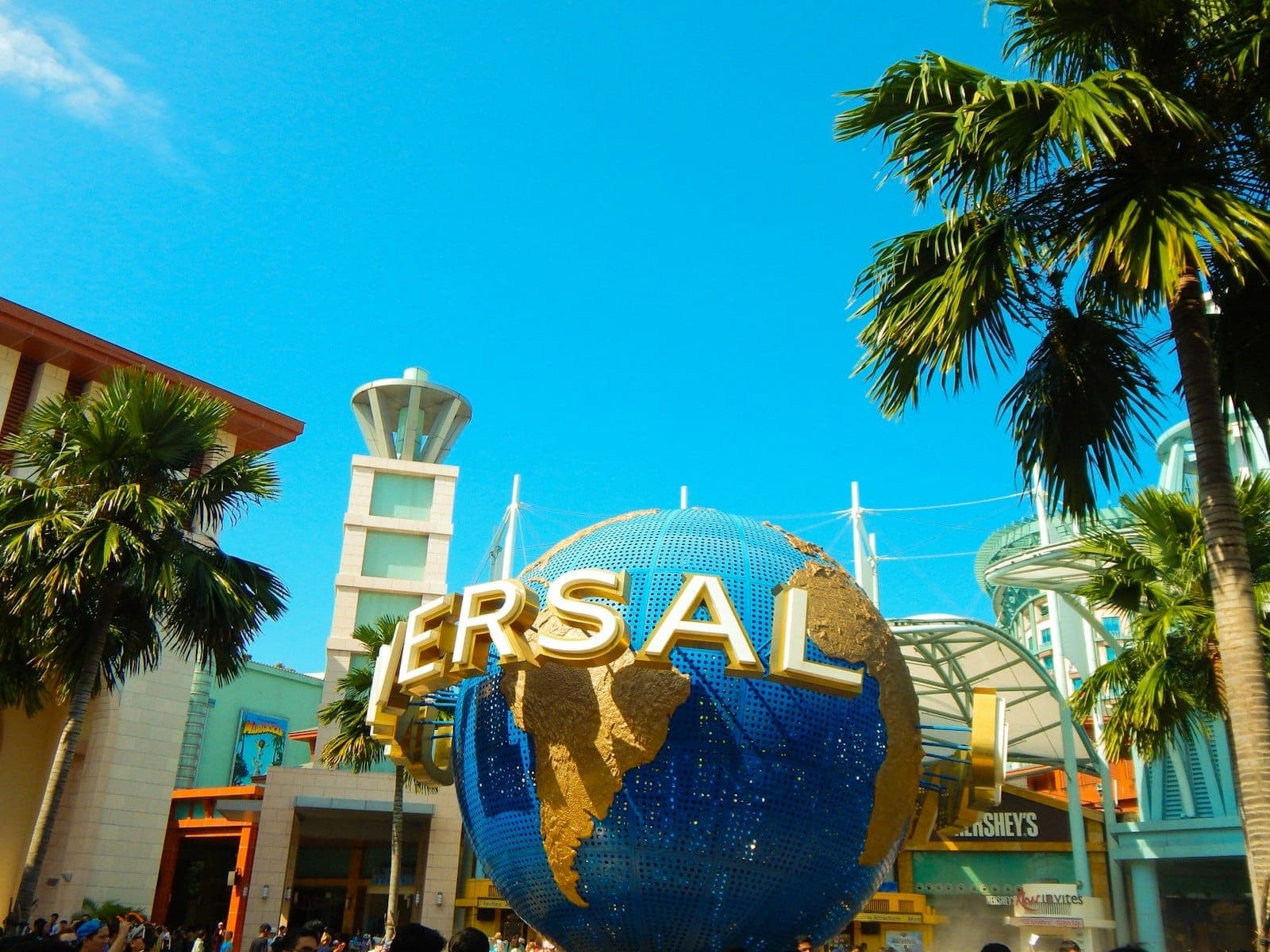 The Universal Studios Singapore entrance features the iconic rotating globe, palm trees, modern buildings, and a clear blue sky.