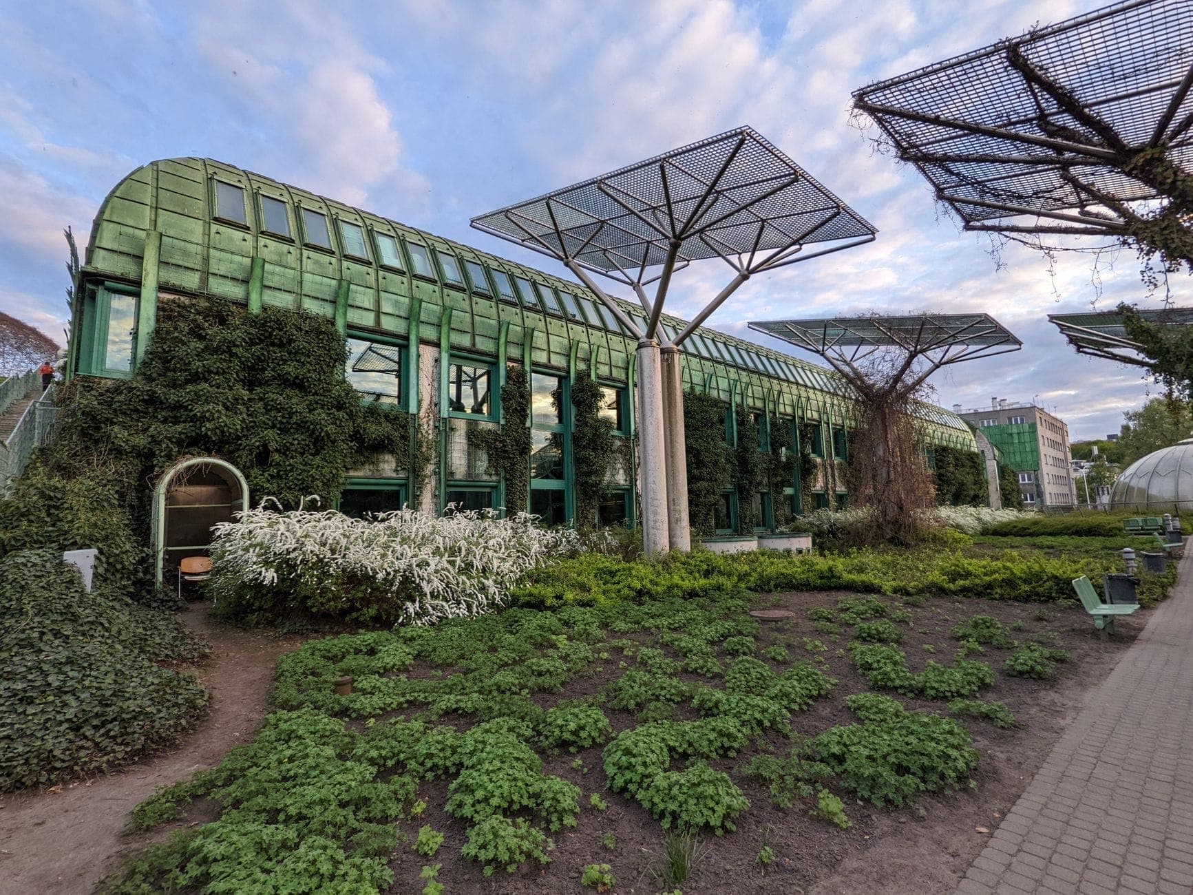 Das üppig begrünte Dach des Bibliotheksgartens der Universität Warschau mit Glasarchitektur, markanten Metallkonstruktionen, Gartenbeeten und Wegen unter einem lebhaften Himmel.