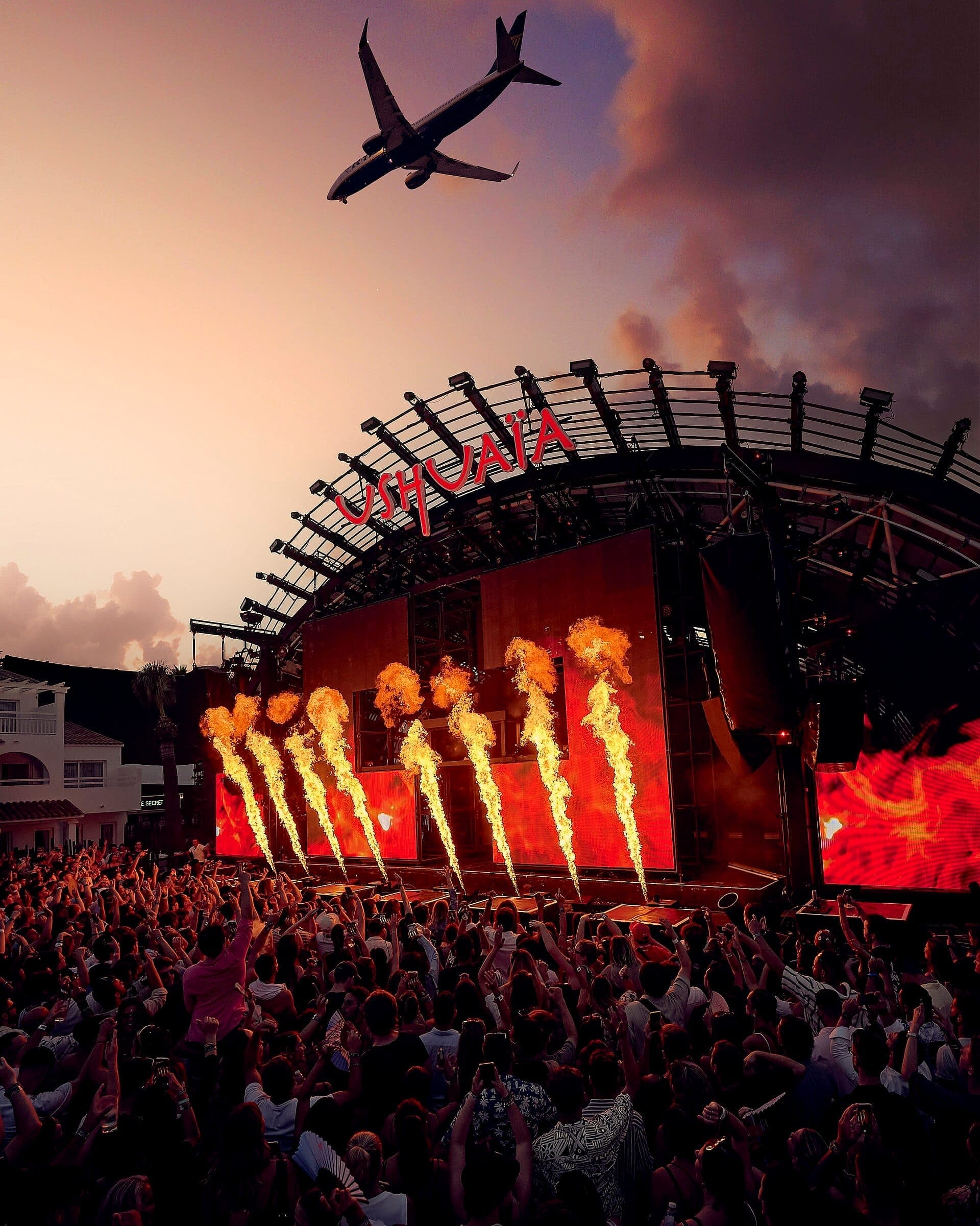 Large crowd at Ushuaïa Ibiza Beach Hotel enjoying a sunset stage show with fire effects and plane flying overhead.