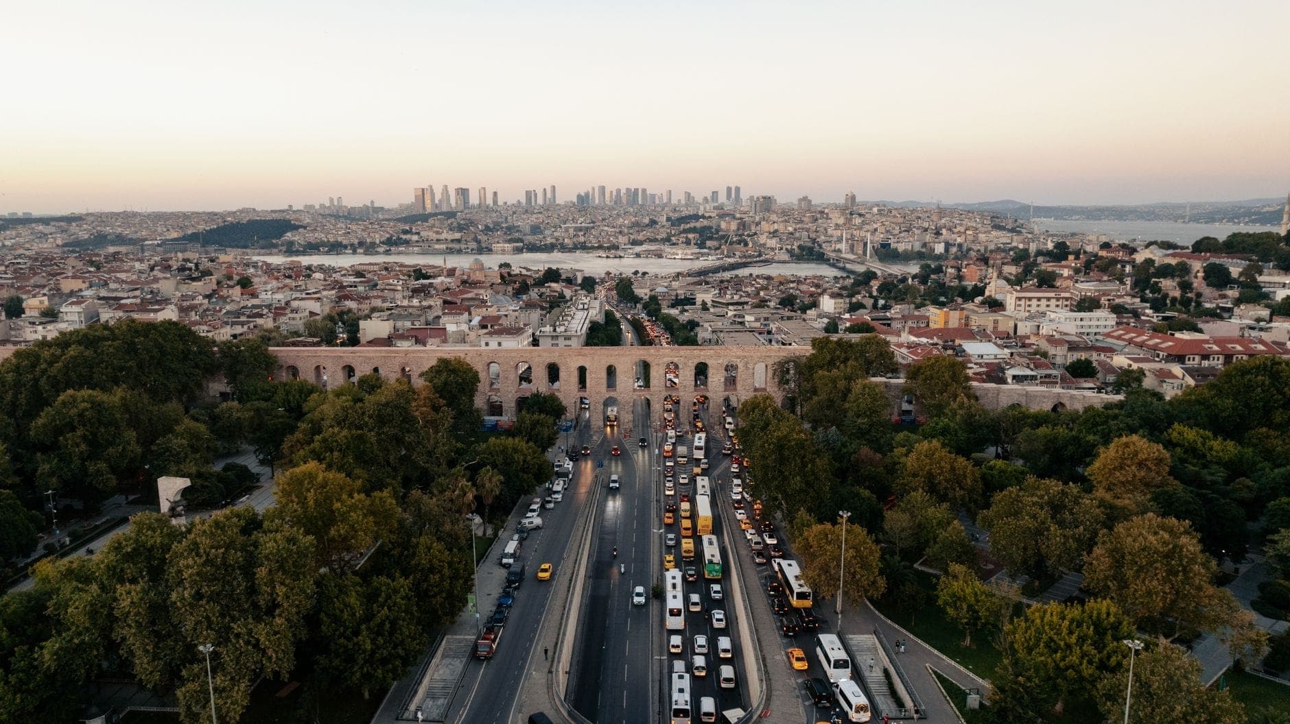 Luftaufnahme des Valens-Aquädukts, der sich über den Atatürk-Boulevard in Istanbul erstreckt, umgeben von Bäumen, mit der Stadtsilhouette und dem Wasser im Hintergrund bei Sonnenuntergang.