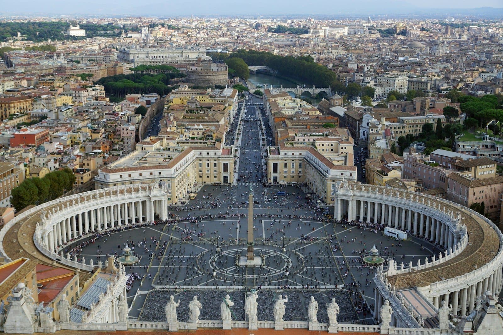 Isang panoramikong tanawin ng Plaza ng San Pedro kasama ang mga iconic na kolonada, mga estatwa, at ang skyline ng Roma sa likod sa ilalim ng maliwanag na langit.
