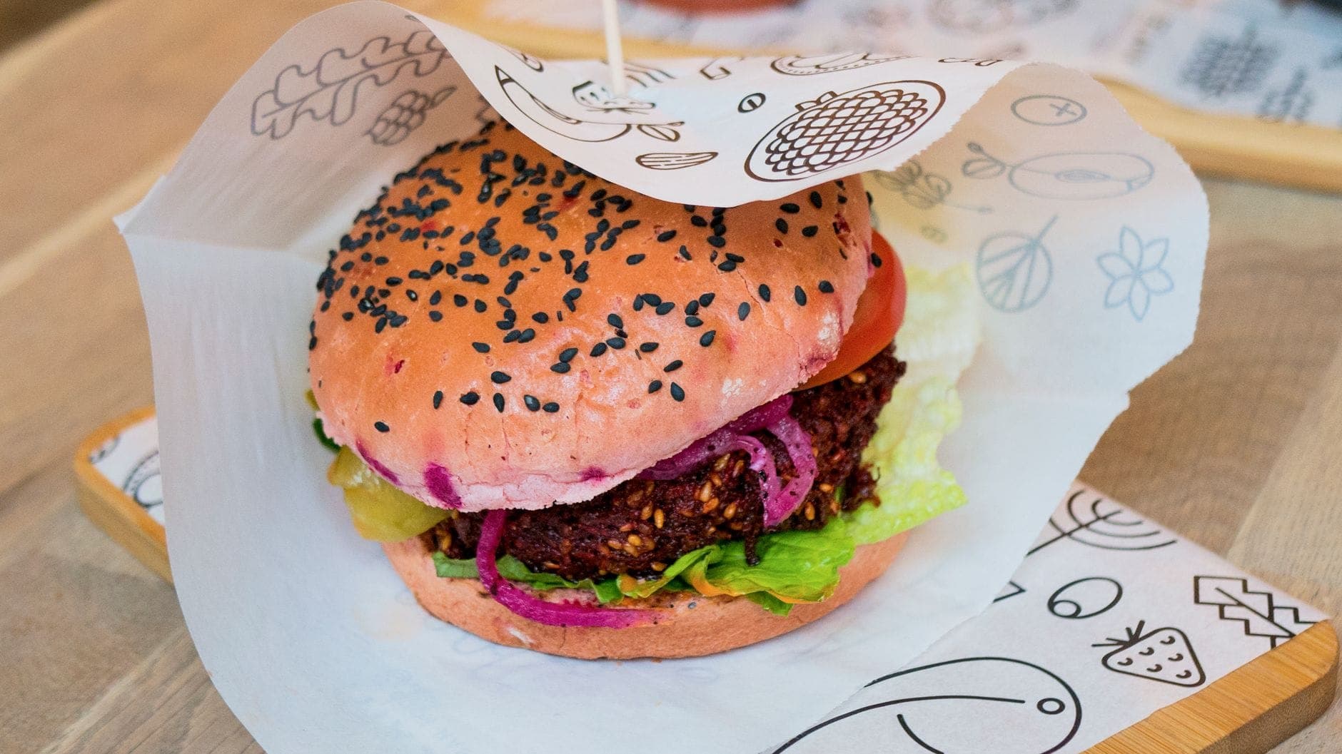 Bánh burger thuần chay đầy màu sắc với vỏ bánh màu hồng, rau xà lách, cà chua và miếng chả, bày trên giấy trang trí trong không gian sáng sủa, thu hút.