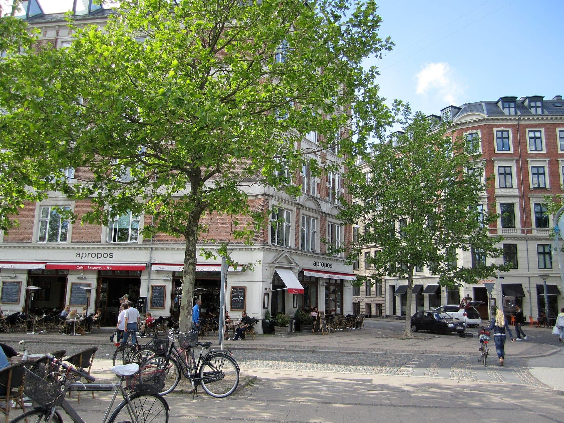 Outdoor-Café-Szene in Vesterbro mit Menschen, die unter grünen Bäumen sitzen, in der Nähe geparkten Fahrrädern und historischen Backsteingebäuden im Hintergrund.