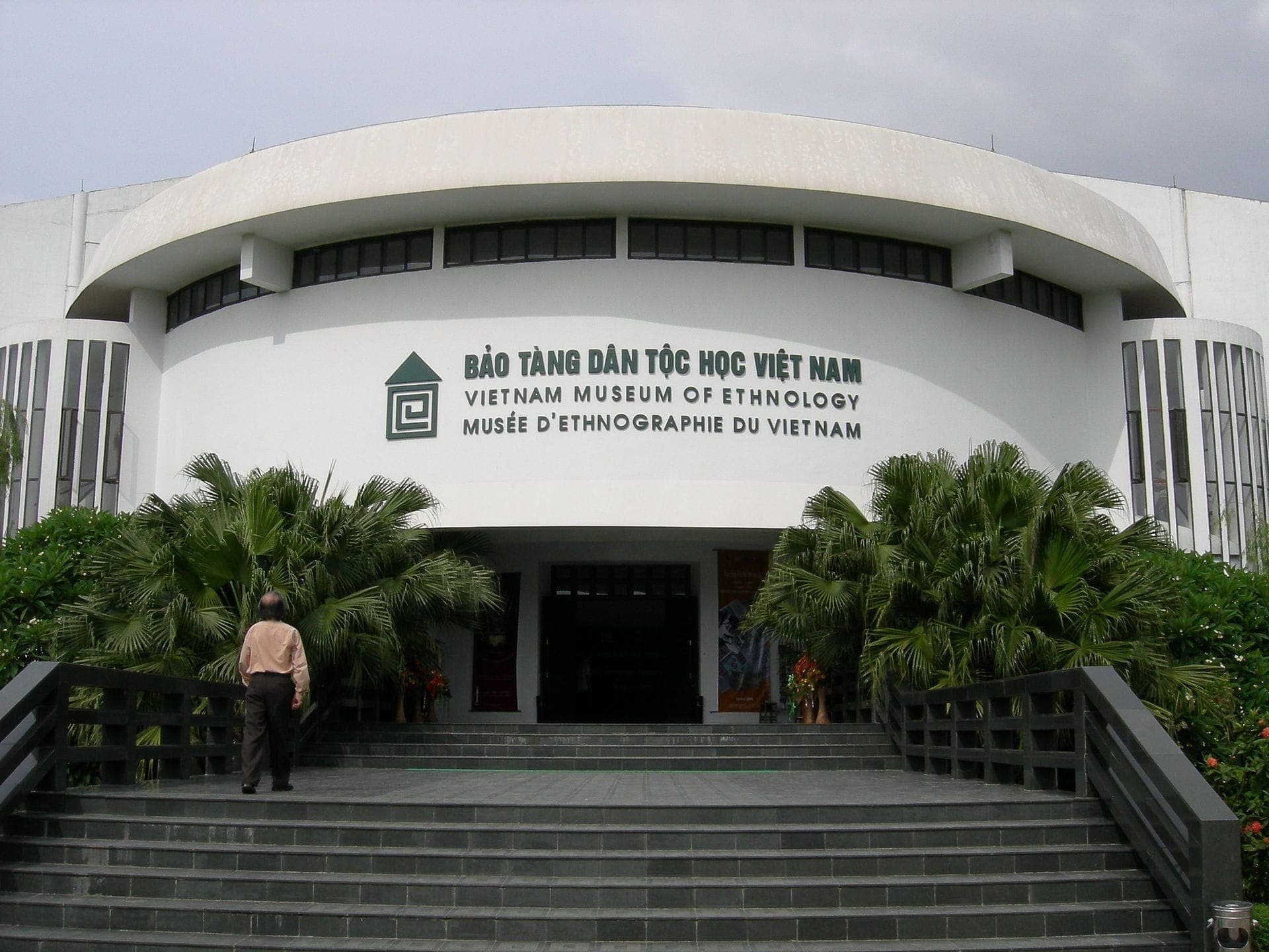 Lối vào chính của Bảo tàng Dân tộc học Việt Nam tại Hà Nội, với những bậc thềm rộng, cây xanh tươi tốt và một người đang tiến về phía cửa ra vào.