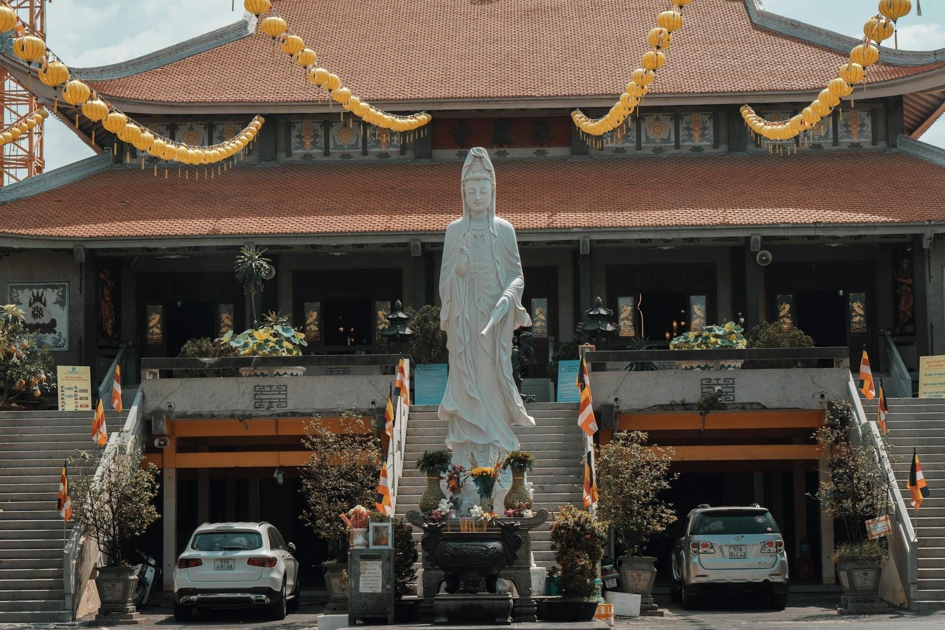 Cảnh mặt tiền chùa Vĩnh Nghiêm với tượng trắng lớn, đèn lồng vàng và xe ô tô đậu dưới ánh sáng ban ngày rực rỡ.