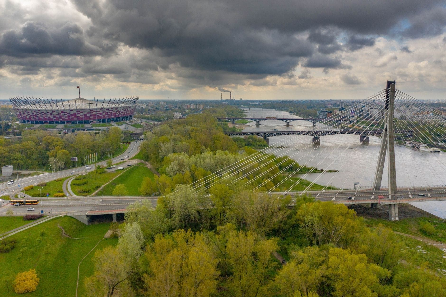 Ein weiter Luftblick auf die Weichselboulevards mit der Świętokrzyski-Brücke und dem Nationalstadion, üppigem Grün, Uferwegen und der Warschauer Skyline unter dramatischen Wolken.