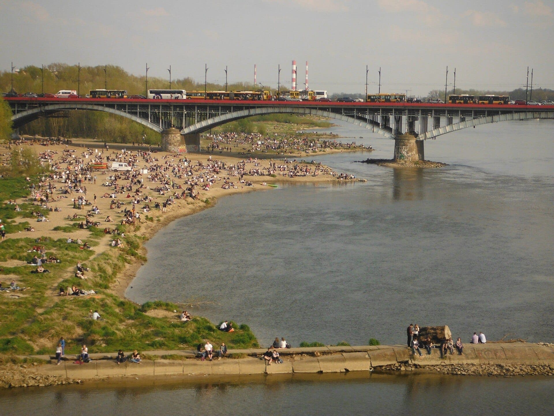 Menschenmengen entspannen sich an den sandigen Ufern des Weichsel-Strandes in Warschau, mit einer Brücke und der Stadtkulisse unter einem teils bewölkten Himmel.