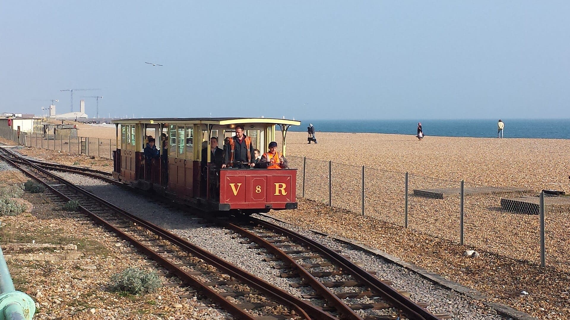 Tàu điện Volk's Electric Railway chạy dọc theo bãi biển sỏi ven biển ở Brighton với hành khách trên tàu và bầu trời xanh trong vắt phía trên.