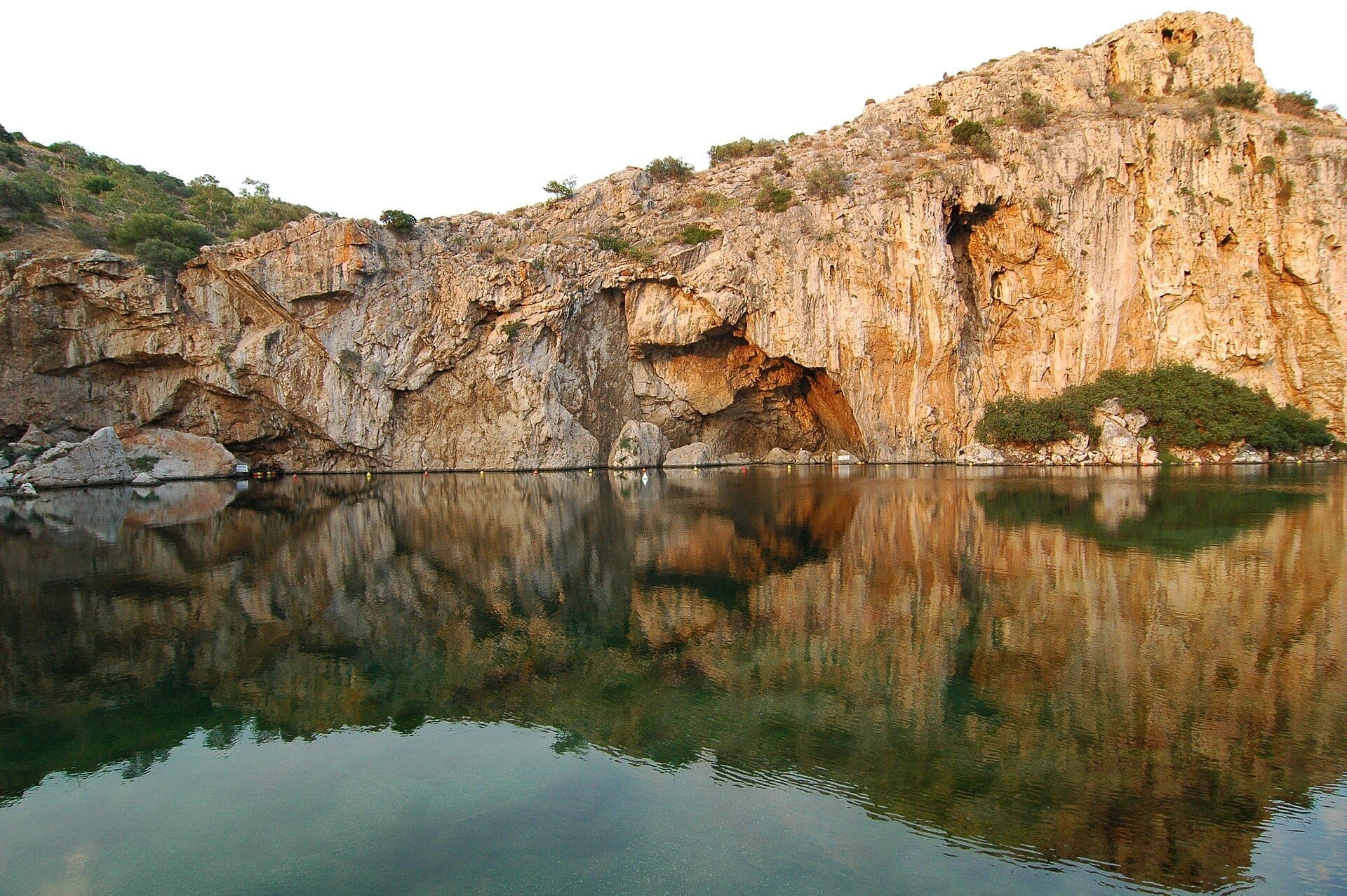 Lake Vouliagmeni with its dramatic rocky cliffs reflected in calm, clear water under a bright sky on the Athenian Riviera.