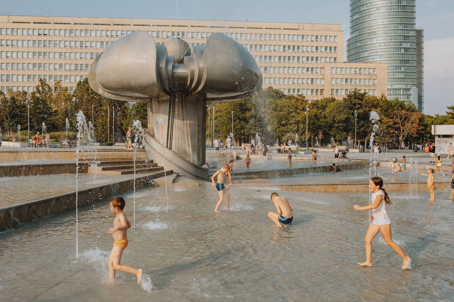 Bambini che giocano e corrono tra fontane moderne in una piazza di Varsavia con una scultura unica e gli edifici della città sullo sfondo.