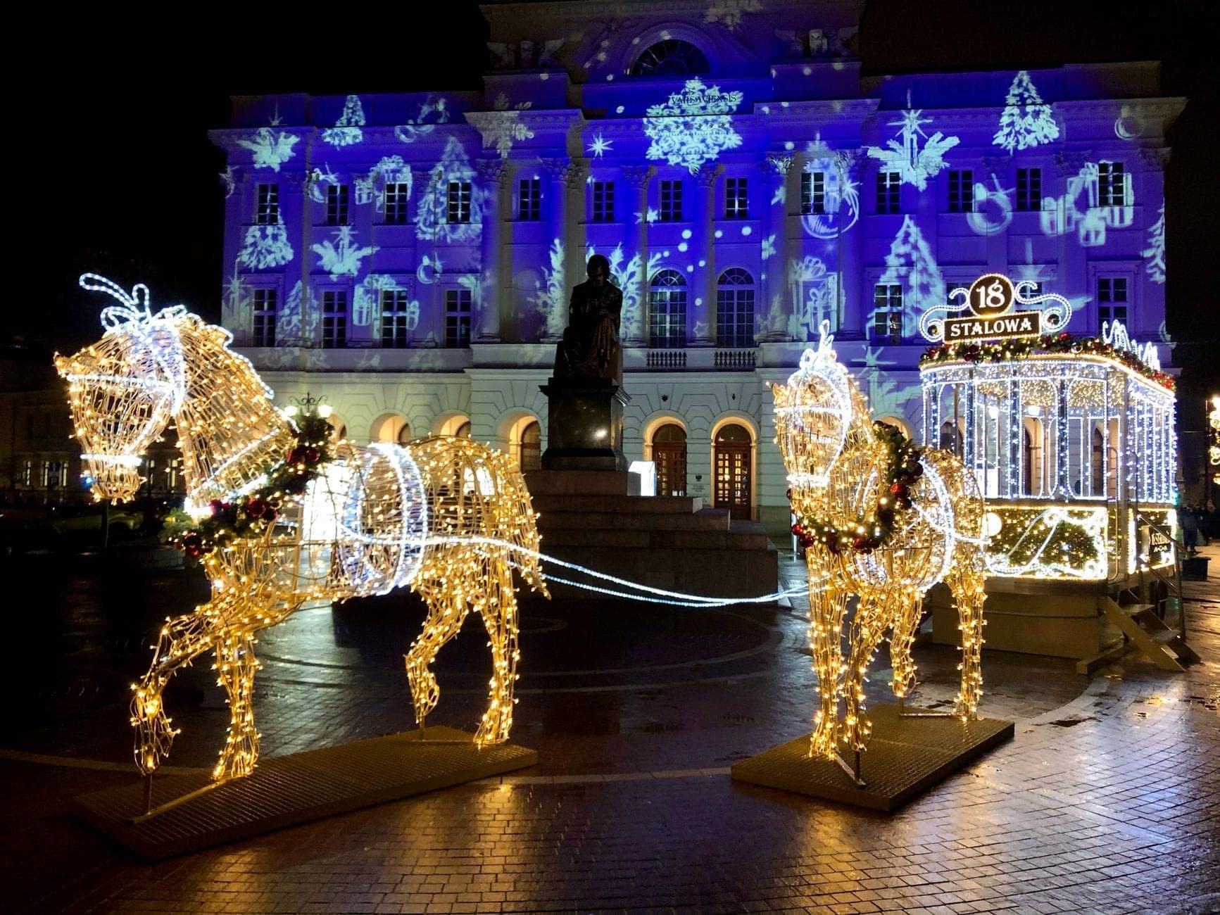 Cảnh đêm tại trung tâm thành phố Warsaw với màn trình diễn đèn Giáng sinh hình ngựa và xe ngựa được chiếu sáng trước một tòa nhà lịch sử nguy nga, cùng các hình chiếu và trang trí lễ hội.