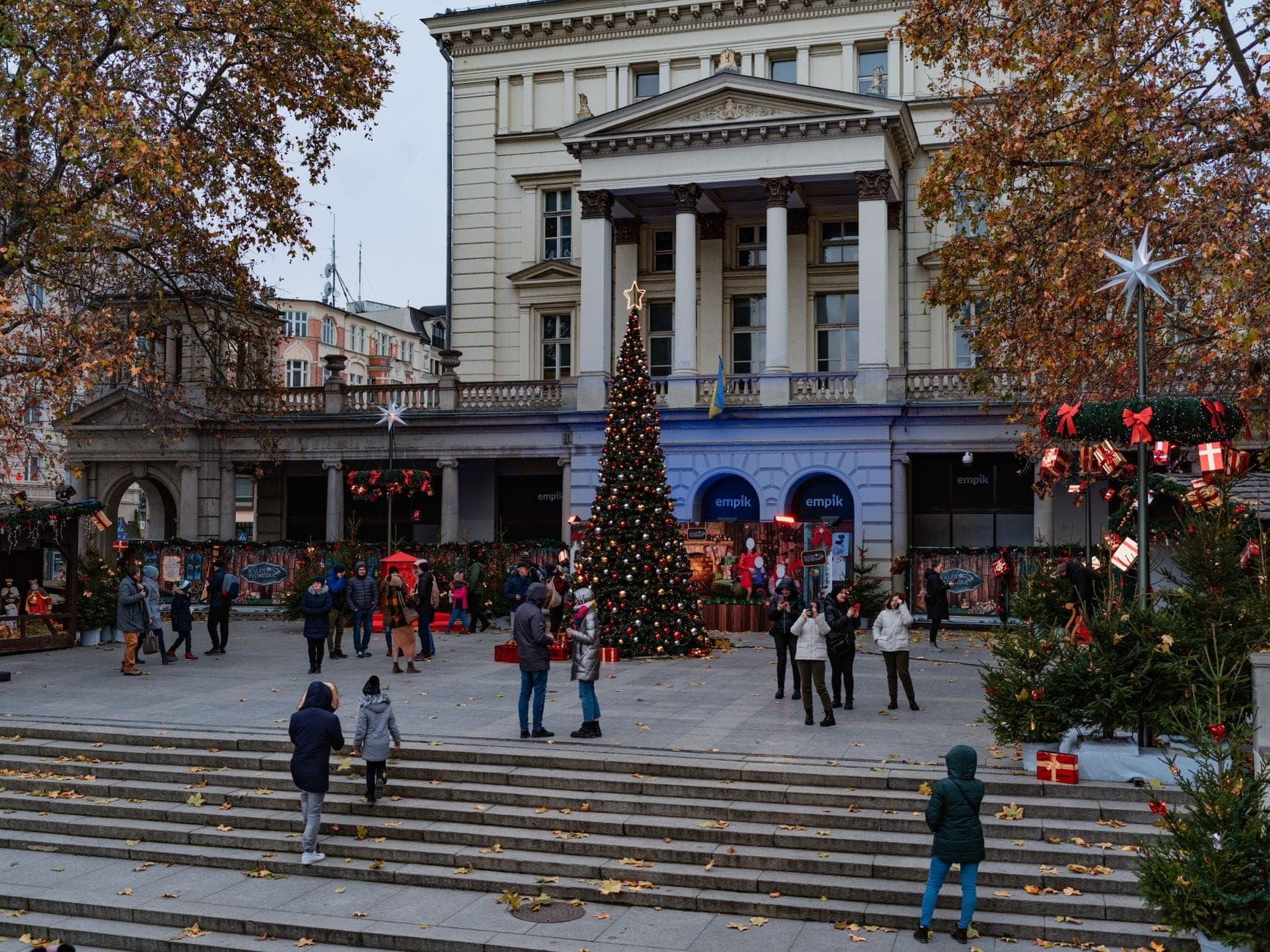 Menschen genießen den Warschauer Weihnachtsmarkt vor einem imposanten neoklassizistischen Gebäude mit einem großen geschmückten Weihnachtsbaum und festlichen Marktständen auf einem Außenplatz.