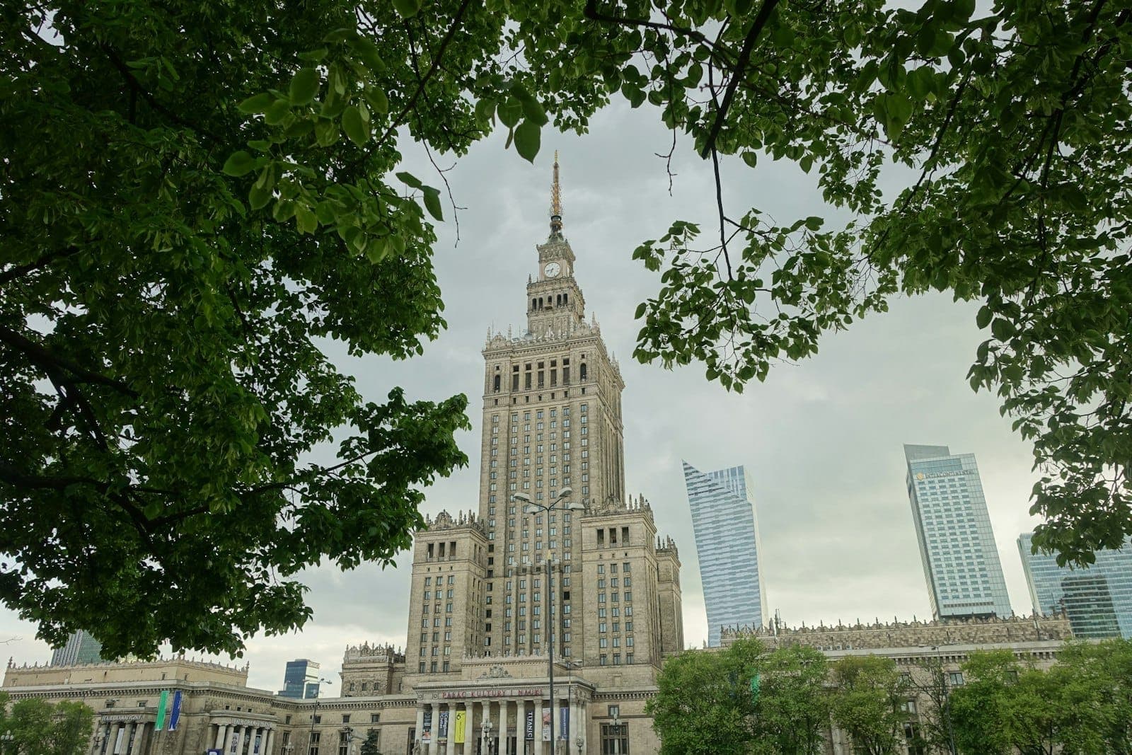 Iconic view of Warsaw’s city center featuring the Palace of Culture and Science framed by leafy green trees, with modern skyscrapers in the background.