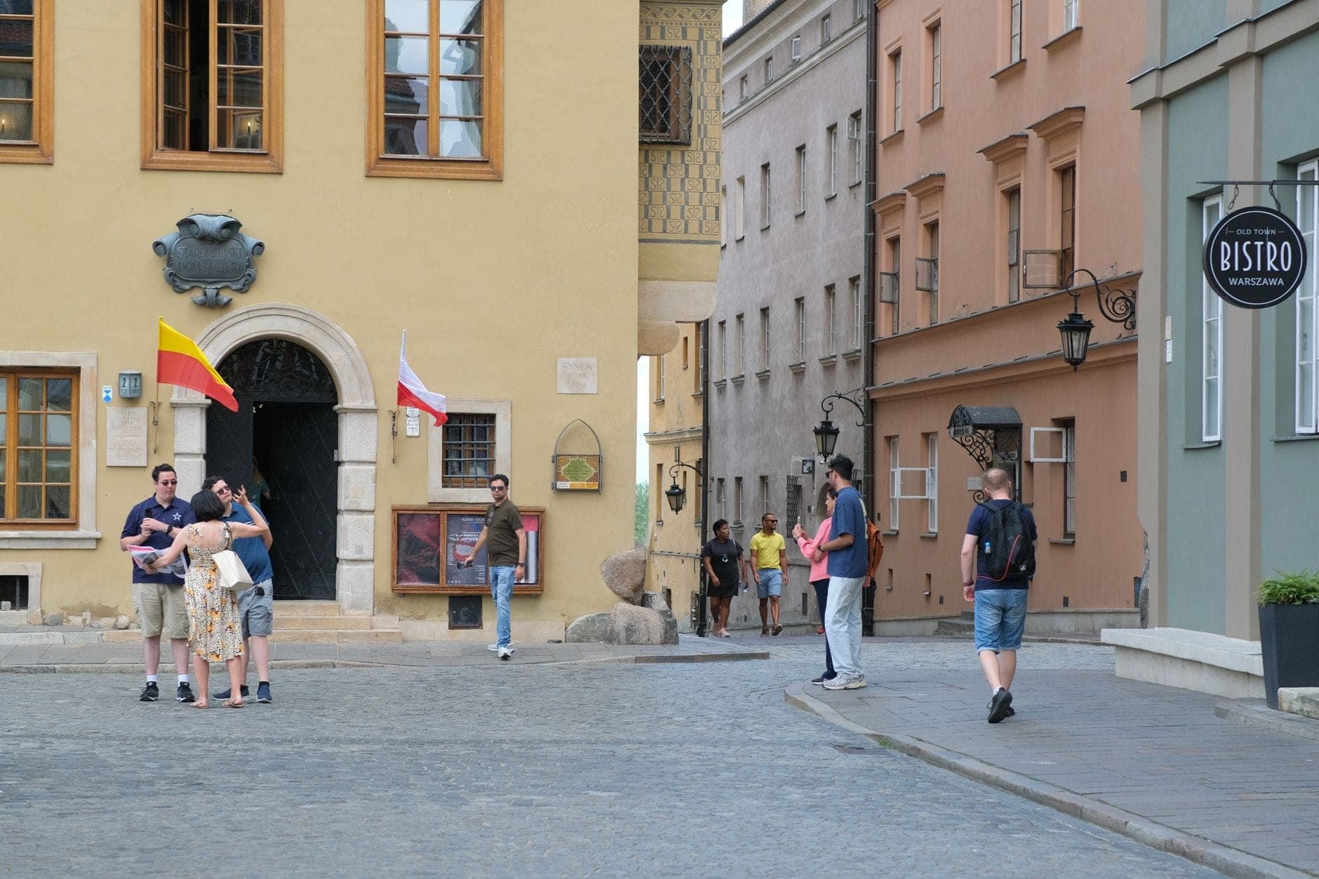 Ludzie spacerujący i rozmawiający na brukowanej ulicy otoczonej kolorowymi zabytkowymi kamienicami i szyldem bistro w Starym Mieście w Warszawie.