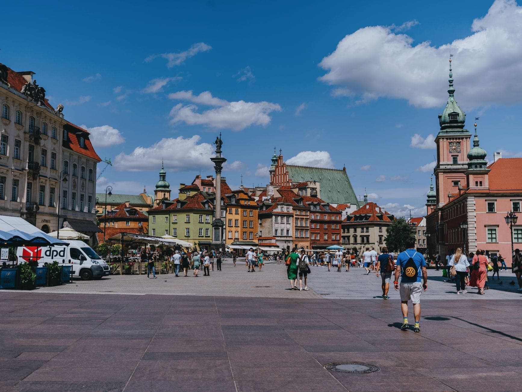 Słoneczna letnia scena Starego Miasta w Warszawie i Placu Zamkowego z przechadzającymi się ludźmi, kolorowymi historycznymi budynkami i błękitnym niebem z chmurami.