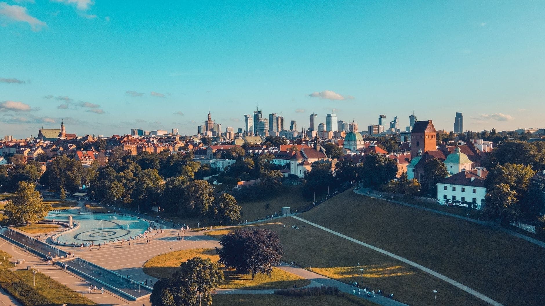 Sommerliche Luftaufnahme von Warschau mit Menschen in einem grünen Park und der Stadtsilhouette unter strahlend blauem Himmel.