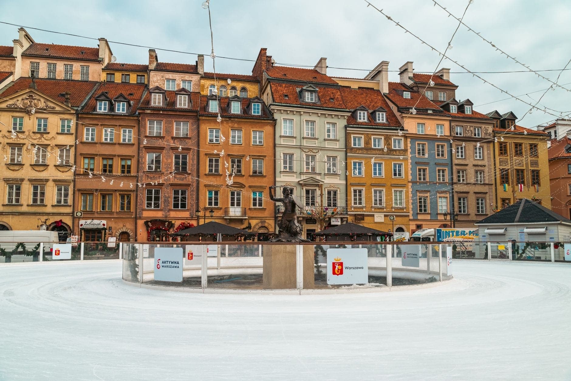 Die bunten Gebäude der Warschauer Altstadt hinter einer zentralen Eislaufbahn, umgeben von Schnee – eine lebhafte Winteratmosphäre im Stadtzentrum.