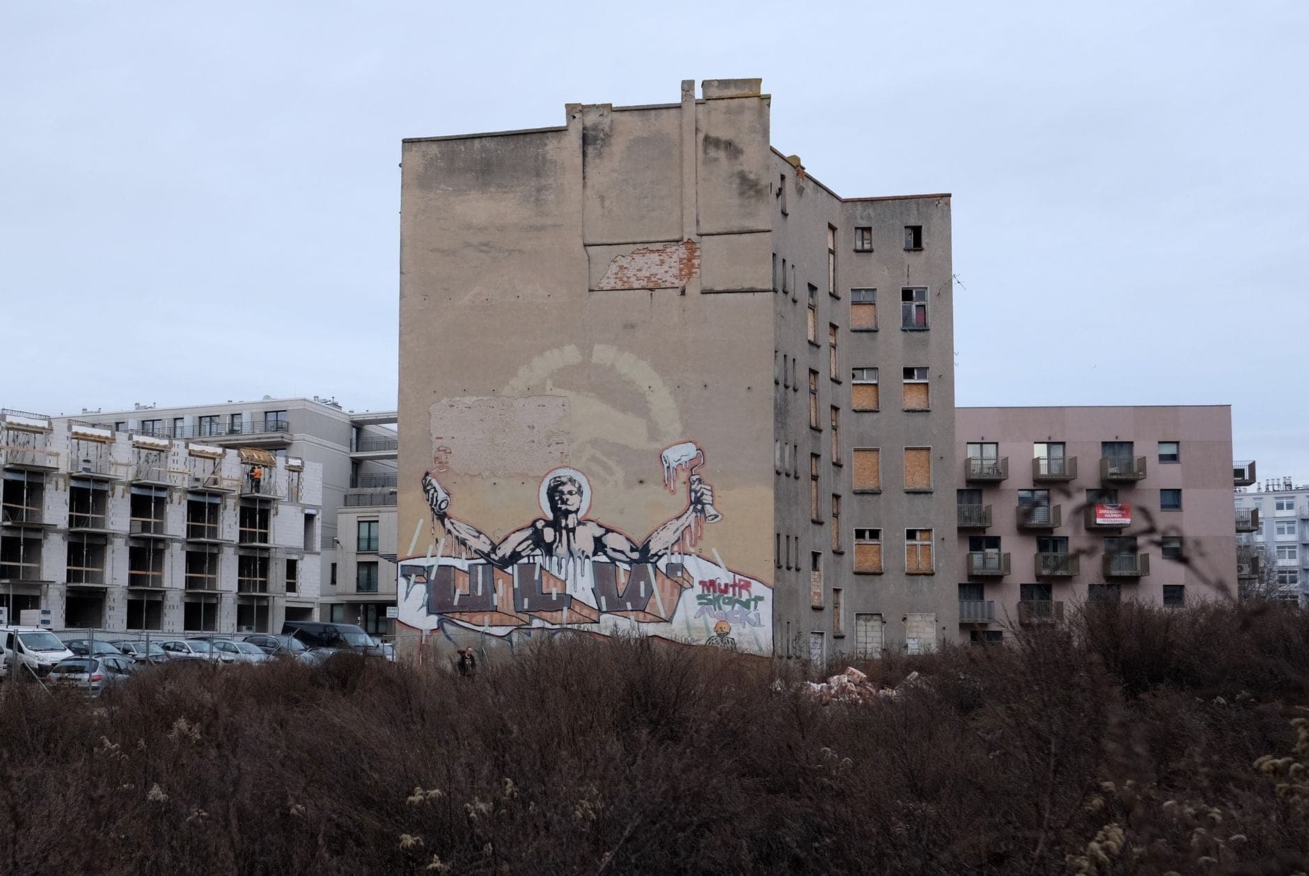 Un lotto vuoto con cespugli incolti, un parcheggio e un palazzo di Varsavia consumato dal tempo con grandi graffiti sul muro—una scena urbana che evoca tracce del passato.