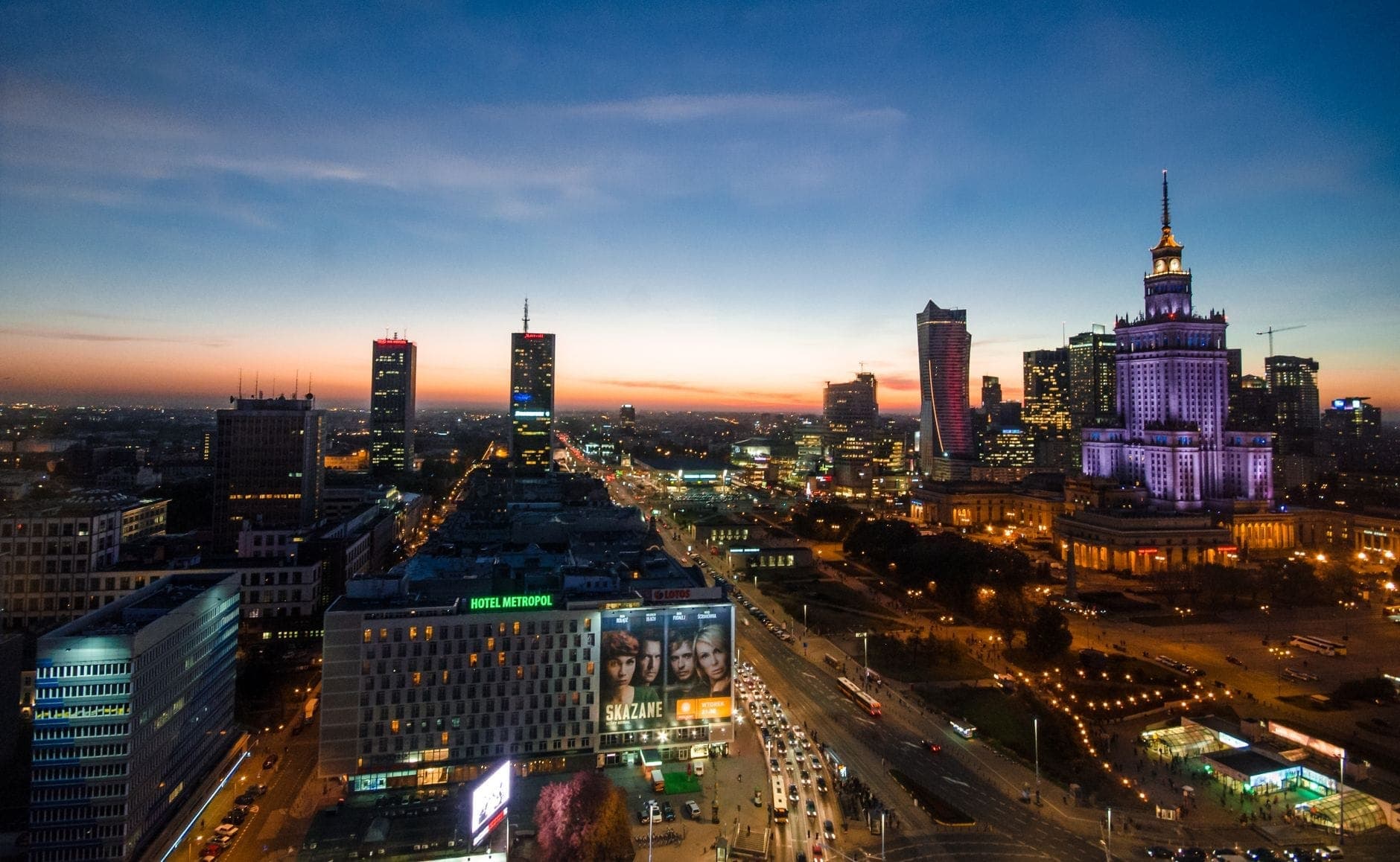 Erhöhte Sonnenuntergangsansicht des Warschauer Stadtzentrums mit modernen Wolkenkratzern und dem beleuchteten Palast der Kultur und Wissenschaft über den belebten Straßen.