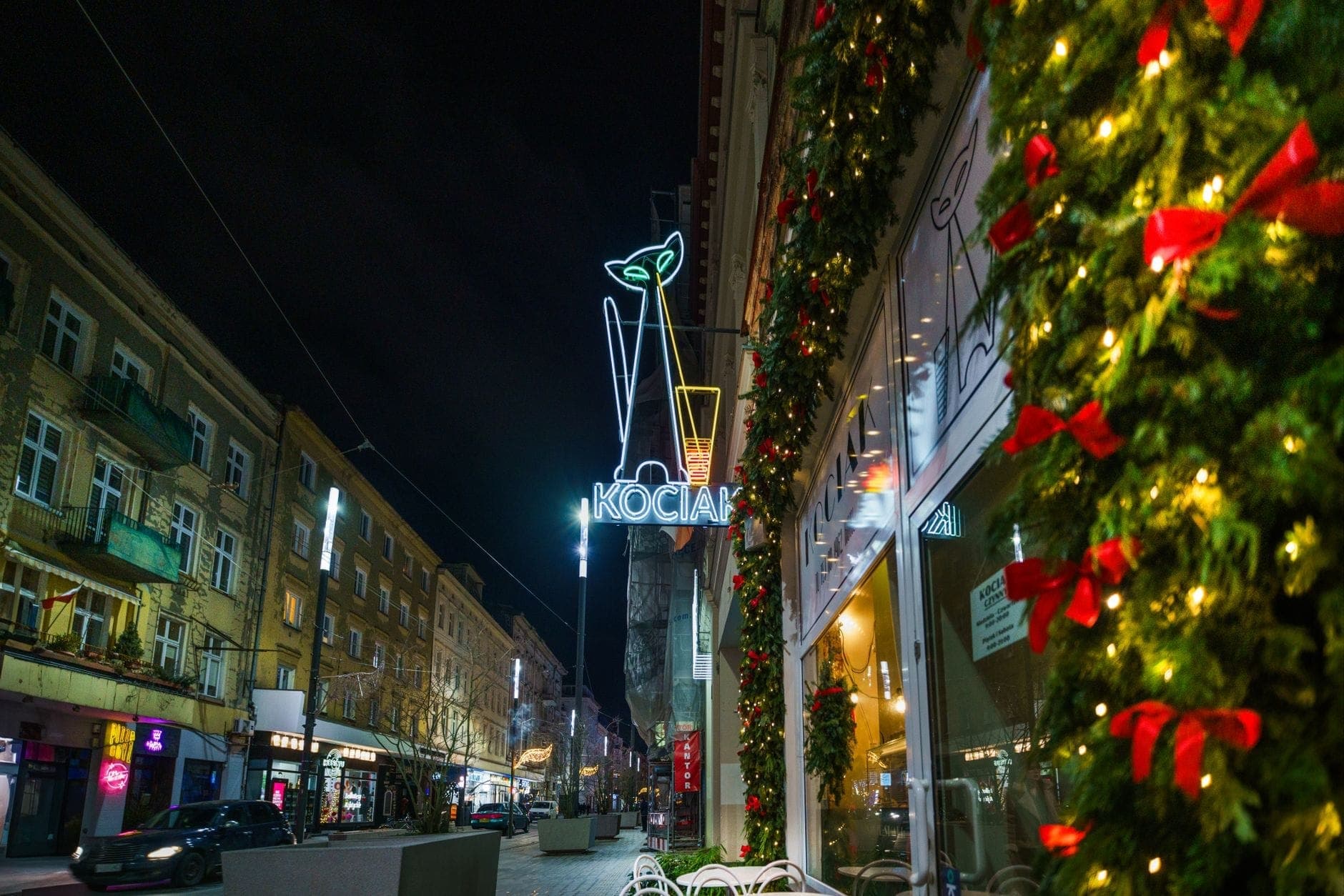 Nächtliche Ansicht einer Straße in Warschau mit Feiertagsdekorationen, Neon-Barschild und klassischen Wohngebäuden entlang des Bürgersteigs.