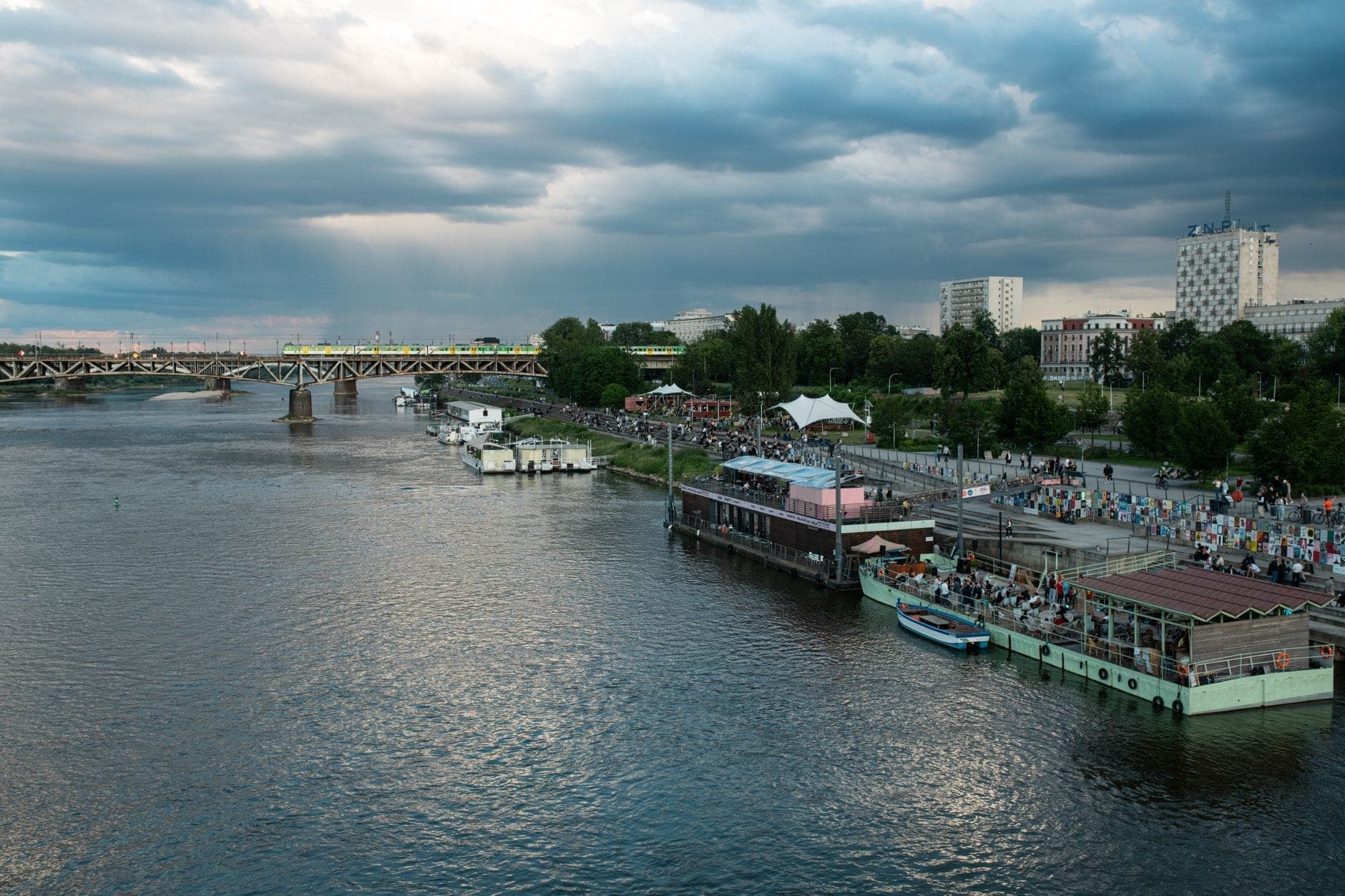 Flussuferszene in Warschau mit schwimmenden Bars, Menschen am Flussufer und Brücken über die Weichsel unter bewölktem Himmel.