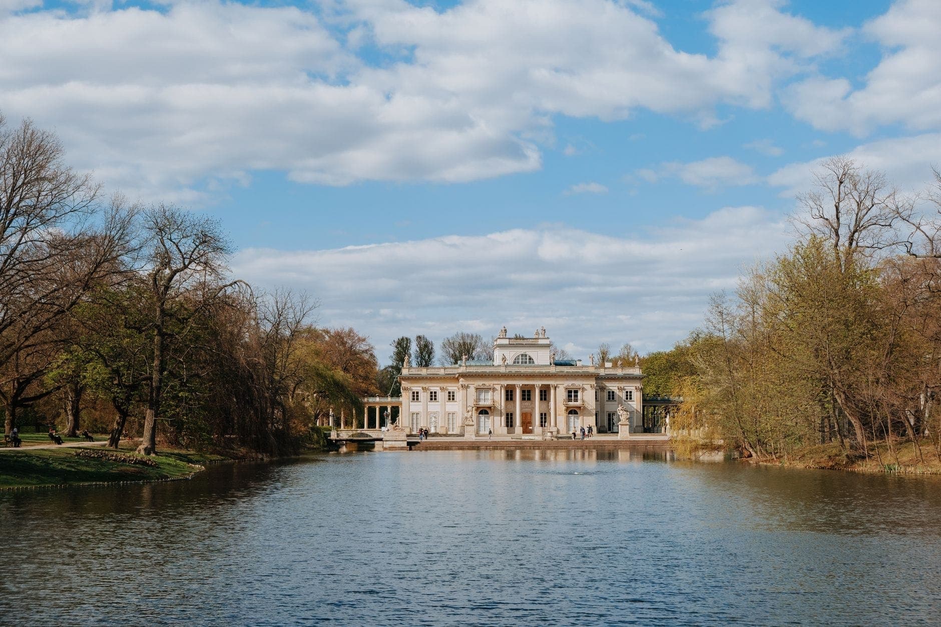 Der Palast auf der Insel im Warschauer Łazienki-Park, umgeben von Bäumen und im stillen See gespiegelt, unter blauem Himmel mit vereinzelten Wolken.