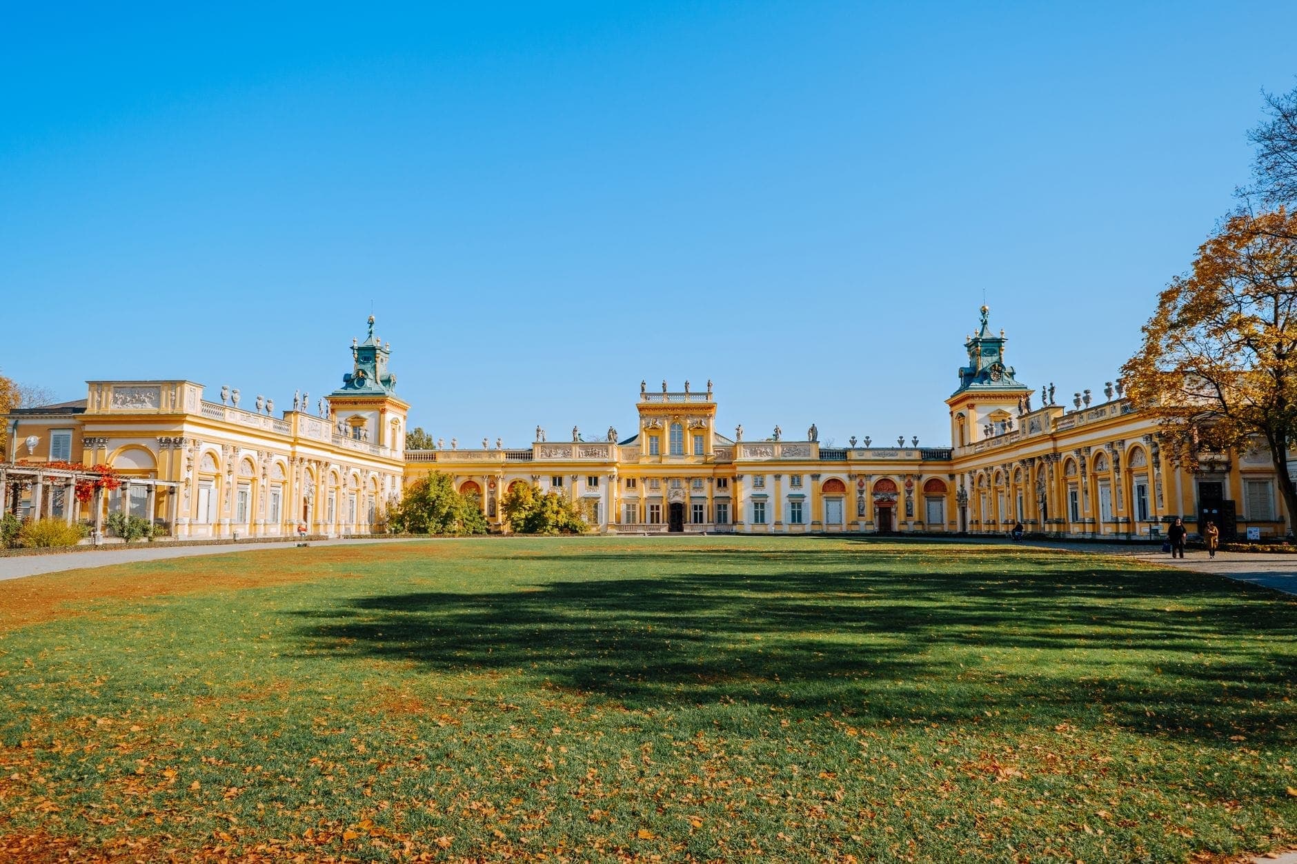 Weitwinkelansicht des Wilanów-Palastes mit seiner prachtvollen gelben Fassade und dem weitläufigen grünen Rasen davor an einem sonnigen Tag.