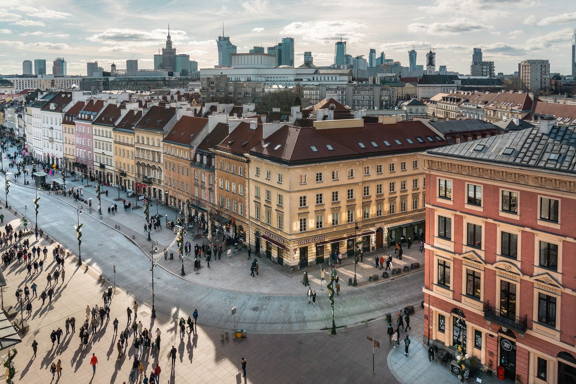 Widok z lotu ptaka na ruchliwą ulicę w centrum Warszawy z tradycyjnymi budynkami, licznymi pieszymi i nowoczesną panoramą miasta w tle.