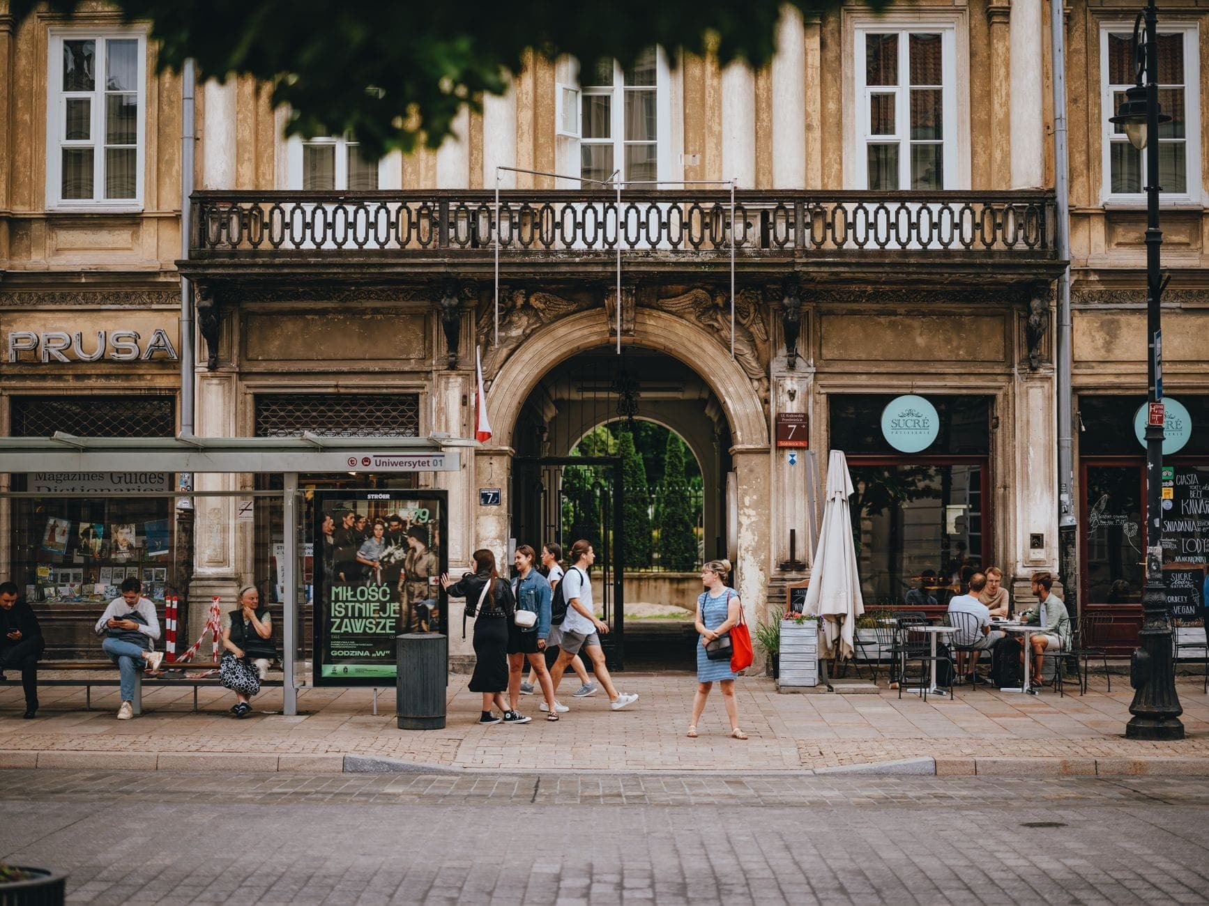 Lebhafte Warschauer Straßenszene mit Menschen vor Geschäften und Cafés vor einem historischen Gebäude – Großstadtatmosphäre pur.