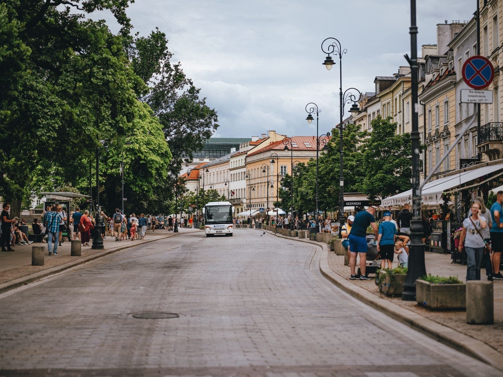 Szeroka, tętniąca życiem ulica piesza w Warszawie z przechodniami, drzewami, historycznymi fasadami, kawiarniami i tramwajem w oddali.