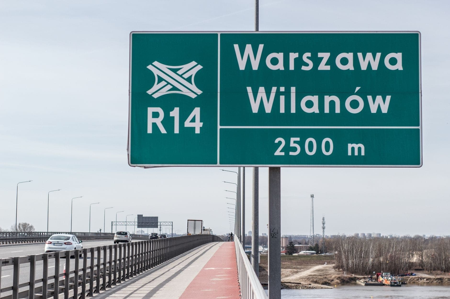 Scena na autostradzie z widocznym znakiem drogowym 'Warszawa Wilanów' i samochodami jadącymi drogą w ciągu dnia.