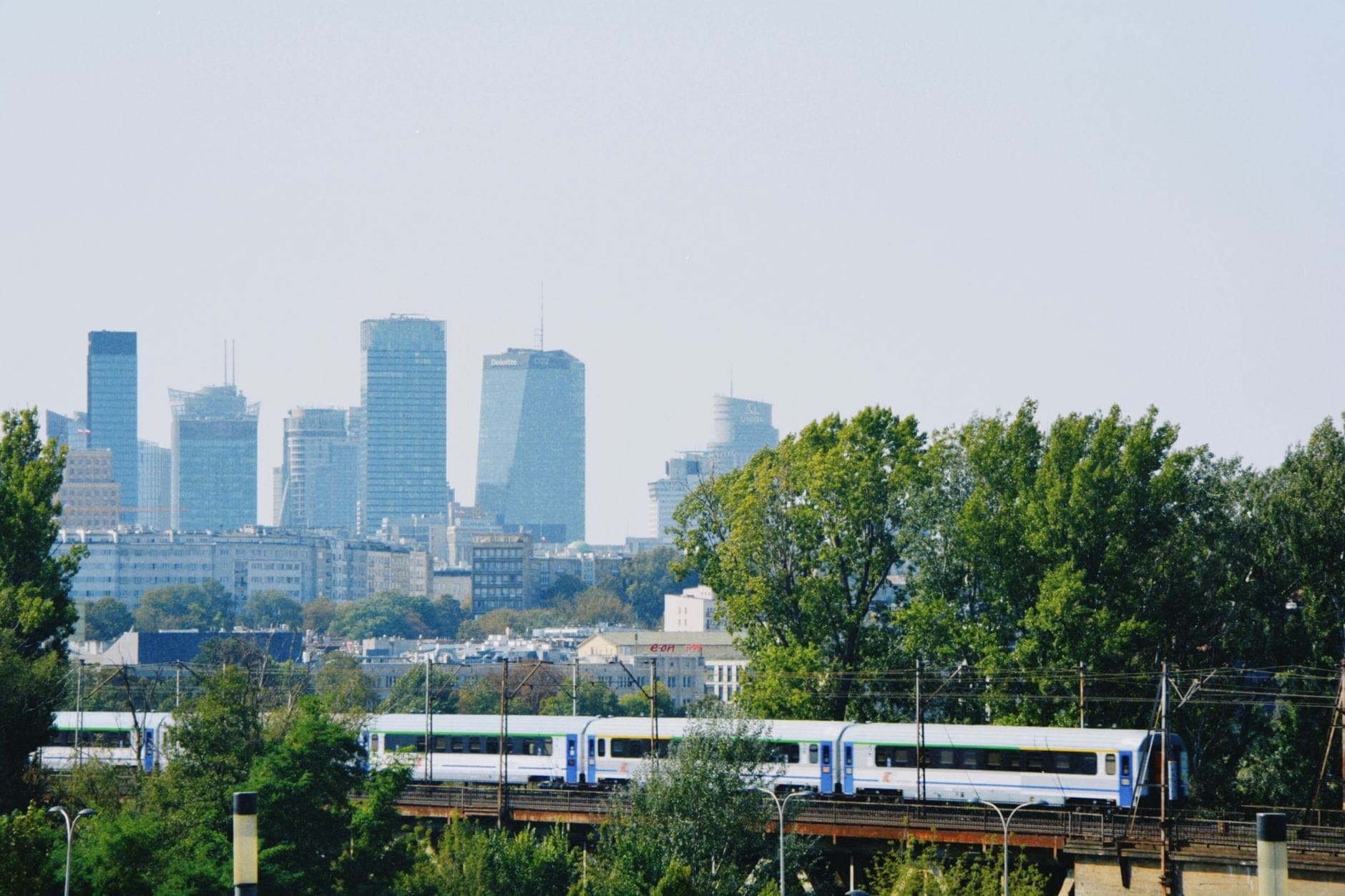 Ein PKP-Intercity-Zug fährt über ein Viadukt, im Hintergrund die Skyline von Warschau, Bäume im Vordergrund, blauer Himmel.
