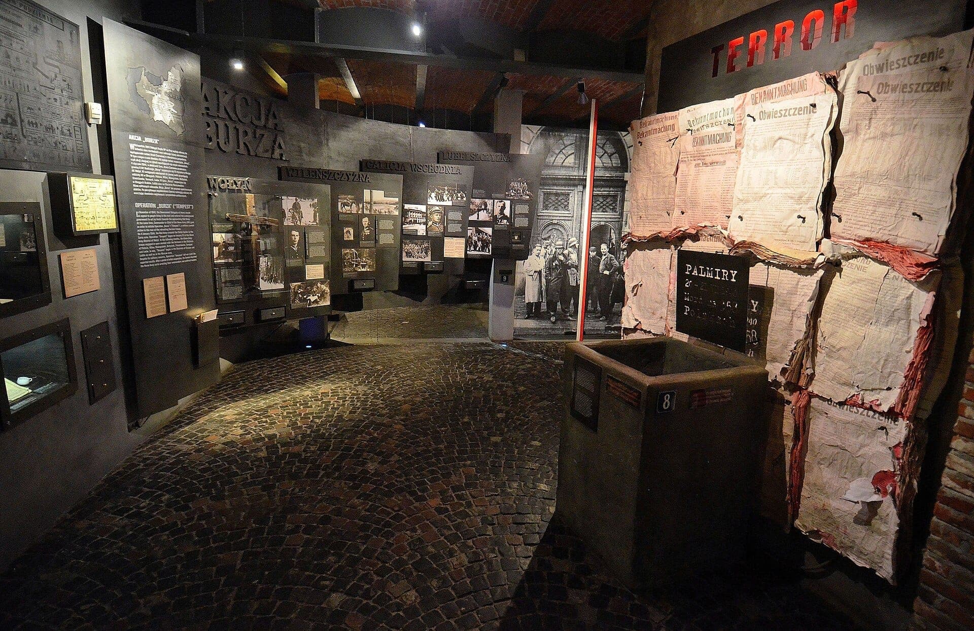 Ausstellungshalle im Museum des Warschauer Aufstands mit historischen Exponaten, Archivfotografien, Kopfsteinpflasterboden und großen Tafeln mit polnischen Dokumenten unter dramatischer Beleuchtung.