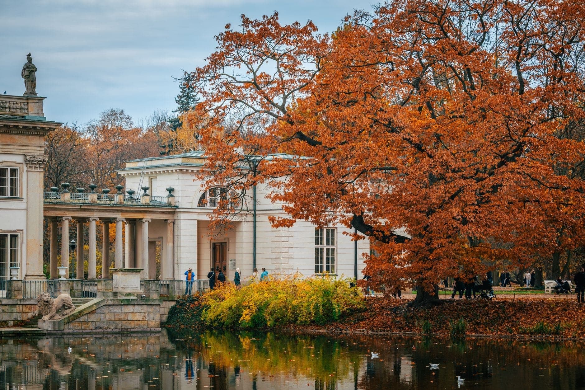 Un grandioso palazzo neoclassico accanto a un lago nel Parco Łazienki, circondato da grandi alberi autunnali e persone che passeggiano all'aperto.