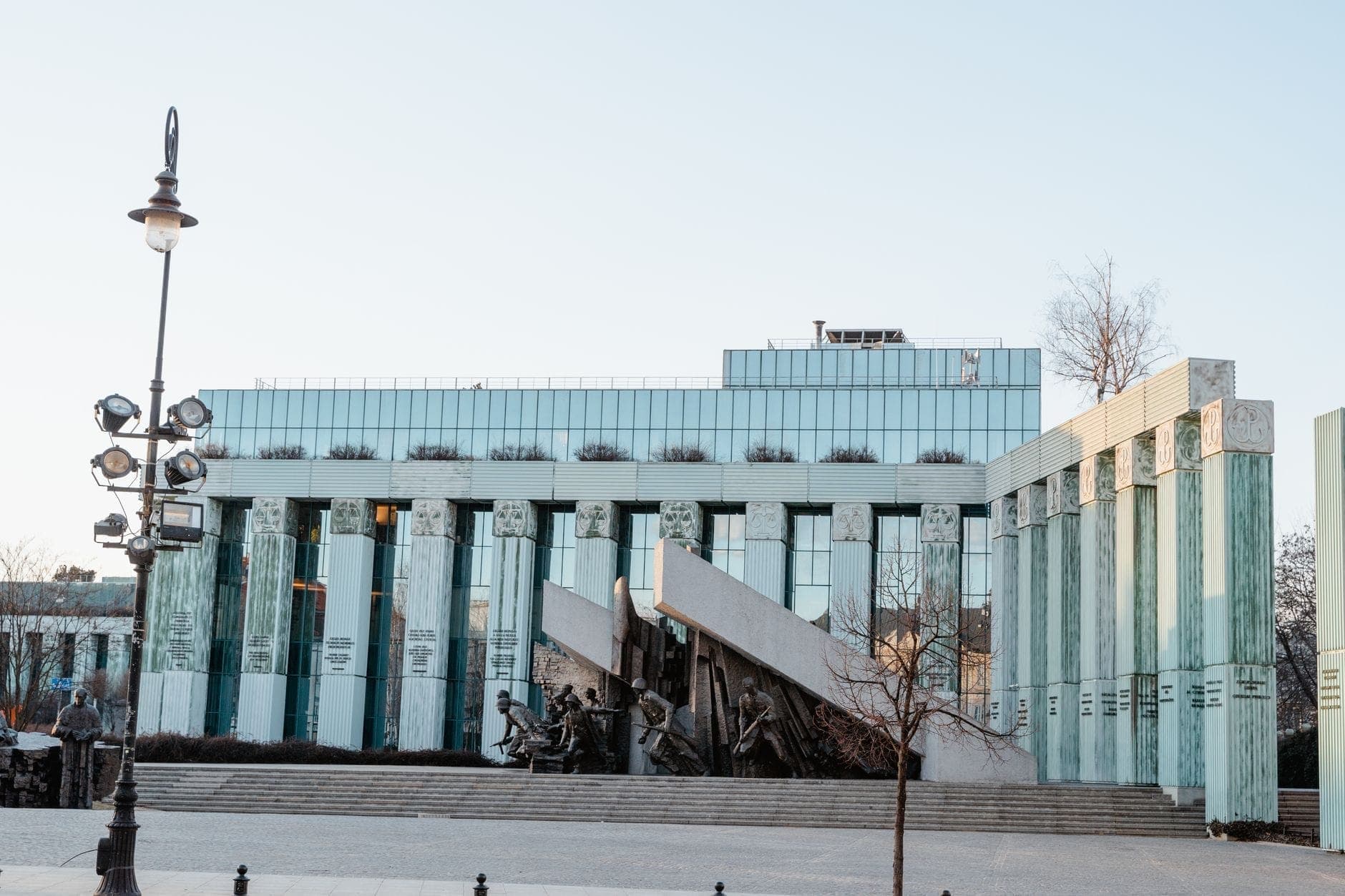 Edificio moderno con alte colonne di vetro verde e il grande Monumento all'Insurrezione di Varsavia in primo piano, che simboleggia la storia accessibile di Varsavia per le famiglie.