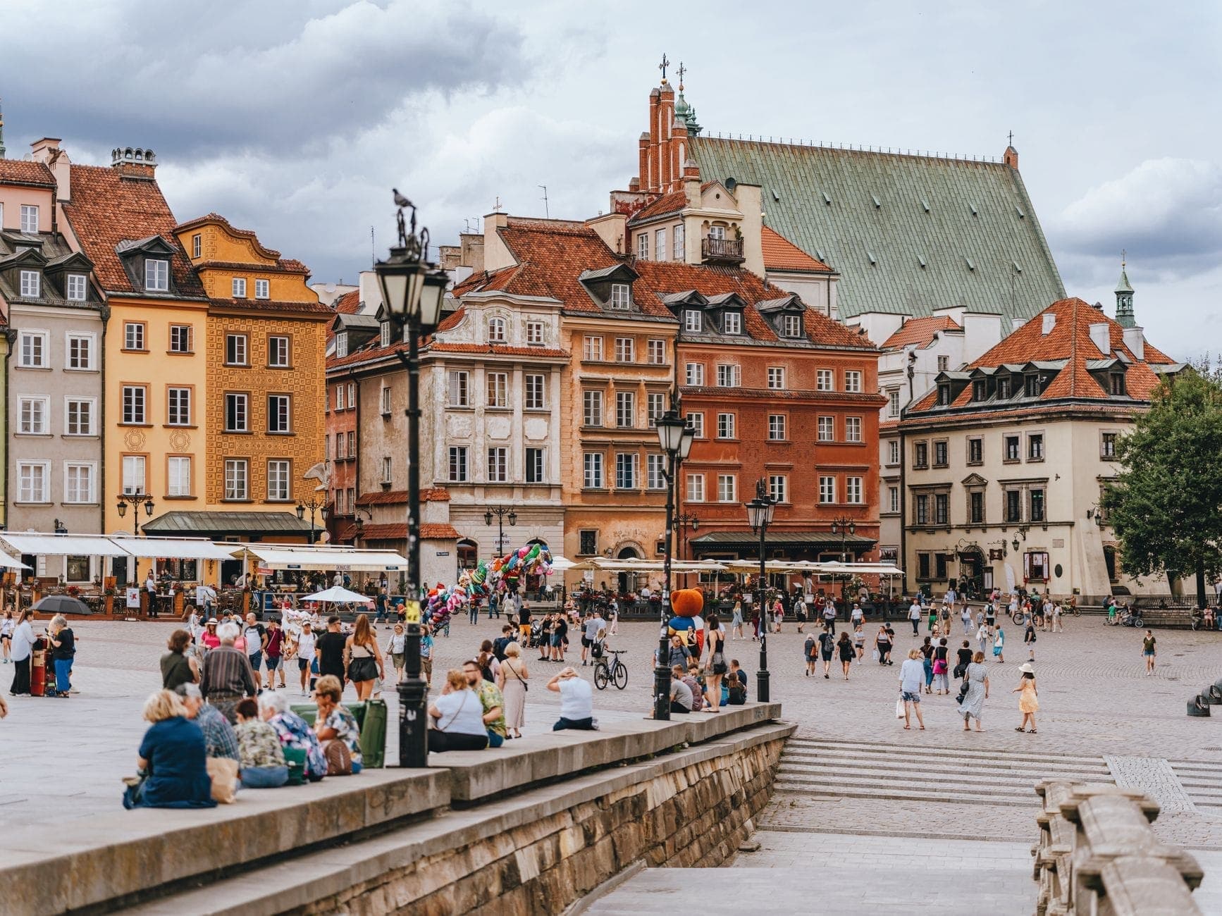 Des foules de personnes sur la Place du Marché de la Vieille Ville reconstruite de Warsaw, avec des façades colorées de style historique et un toit d'église proéminent en arrière-plan.