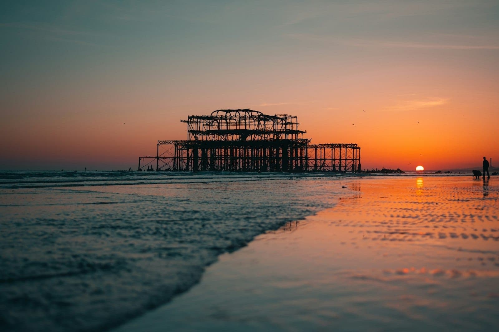 Tàn tích khung xương của West Pier Ruins sừng sững ngoài khơi Brighton lúc hoàng hôn, phản chiếu trên bãi cát ướt và mặt biển yên tĩnh.